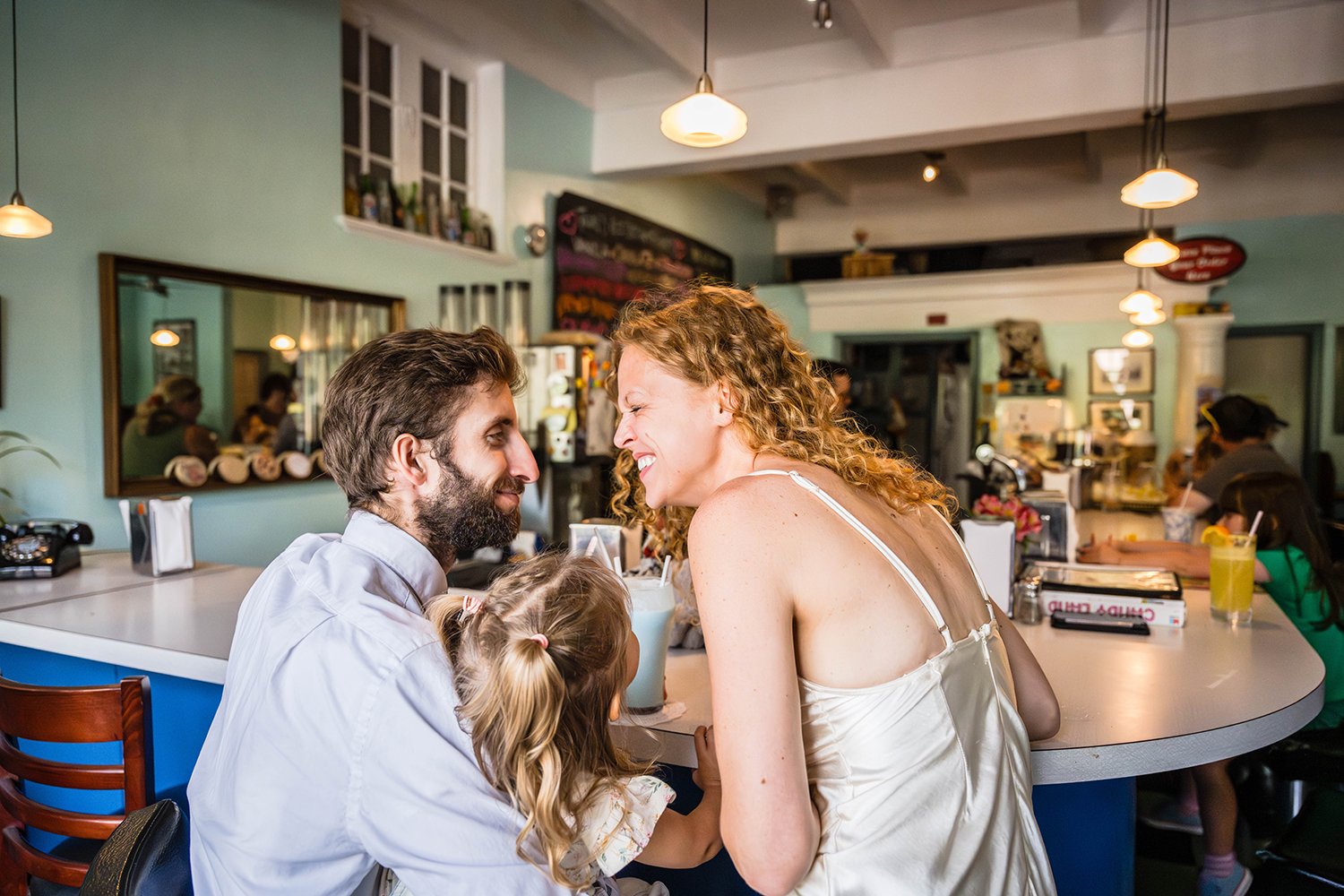 A bride and groom smile at one another widely as their toddler eagerly looks up. The family of three are seated at the bar of Pop’s Ice Cream &amp; Soda Bar  in Grandin Village. 