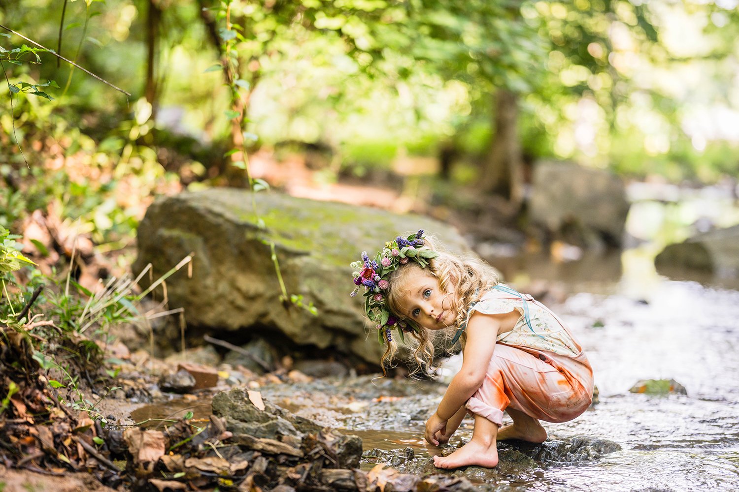 A toddler wearing eco-dyed bib overalls and a flower crown plays in the creek and looks up for a photo at Fishburn Park in Roanoke, Virginia.