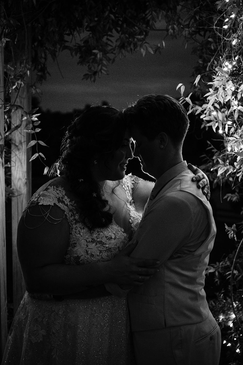 An LGBTQ+ couple embraces one another under a ceremony arch adorned with plants and twinkling lights on their wedding day.