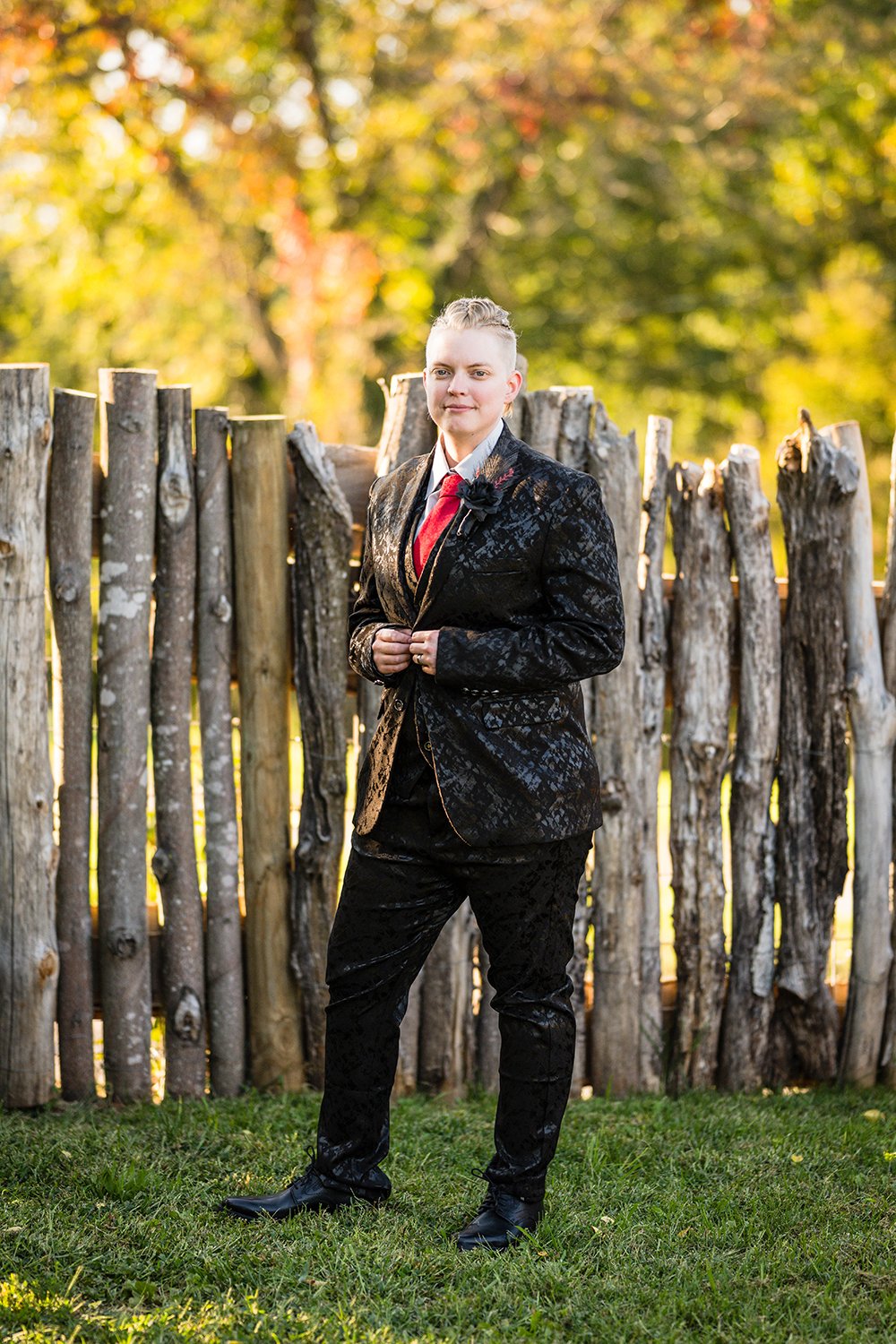A lesbian bride wears a black floral suit and stands against a wooden log fence in the yard during their elopement.