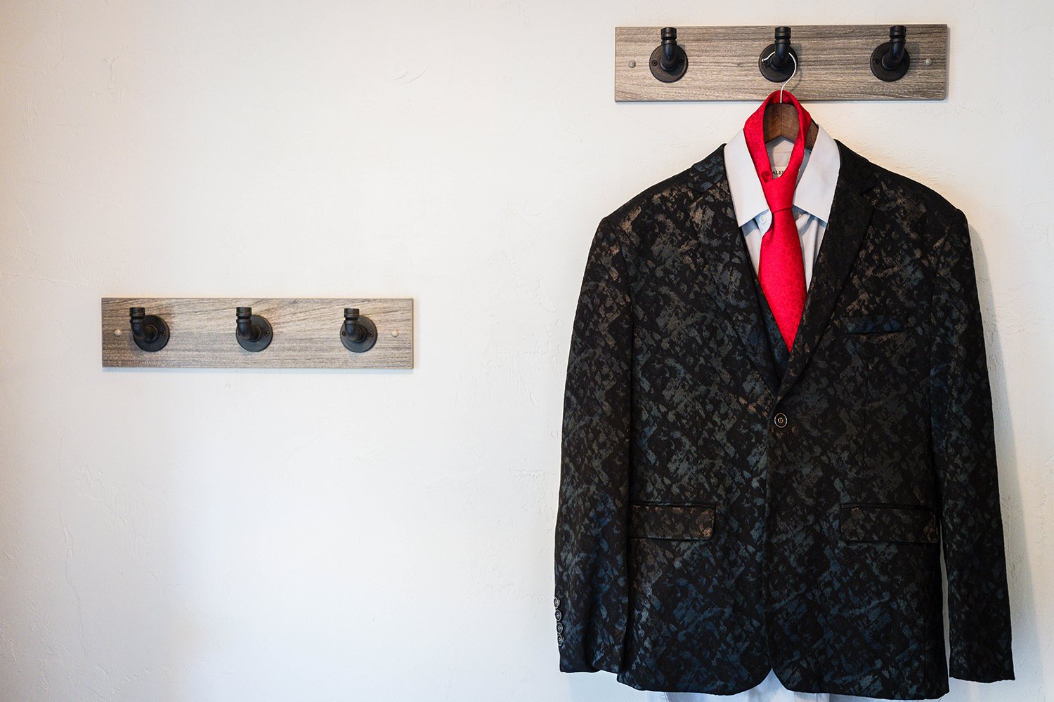 A black floral wedding suit jacket, gray undershirt, and red floral tie hangs on a wooden hanger on a coat rack by the backdoor of a Roanoke Airbnb.
