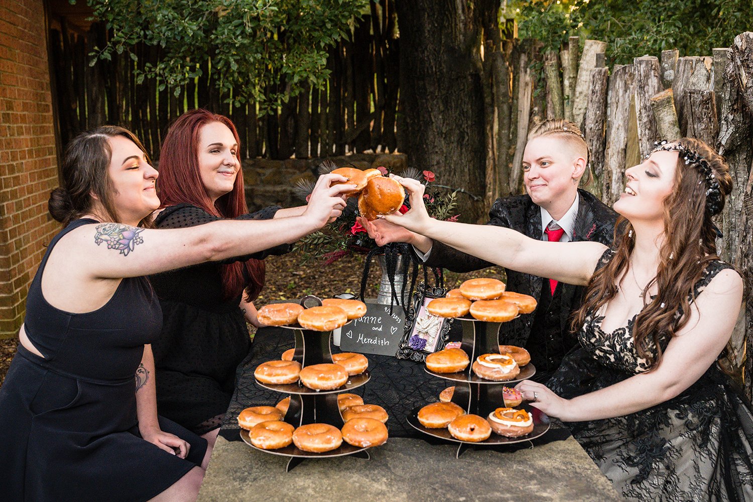 A lesbian wedding couple and their wedding party sit at a stone table and “clink” donuts on their elopement day at a Roanoke Airbnb.