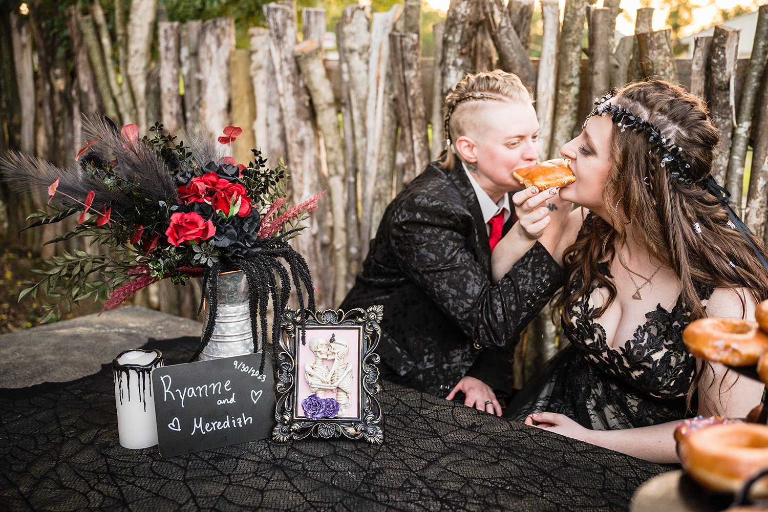 A queer wedding couple intertwine arms and eat donuts while seated at a stone table decorated with black tablecloth during their Roanoke airbnb elopement.