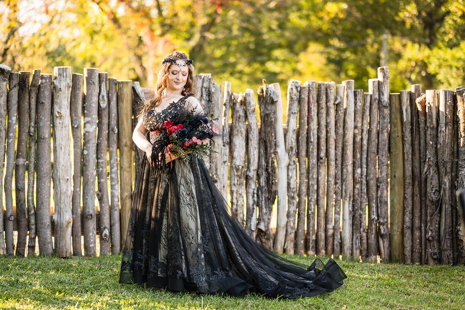 A lesbian marrier wears a black wedding dress and holds a wedding bouquet against a wooden log fence in the yard during their elopement.