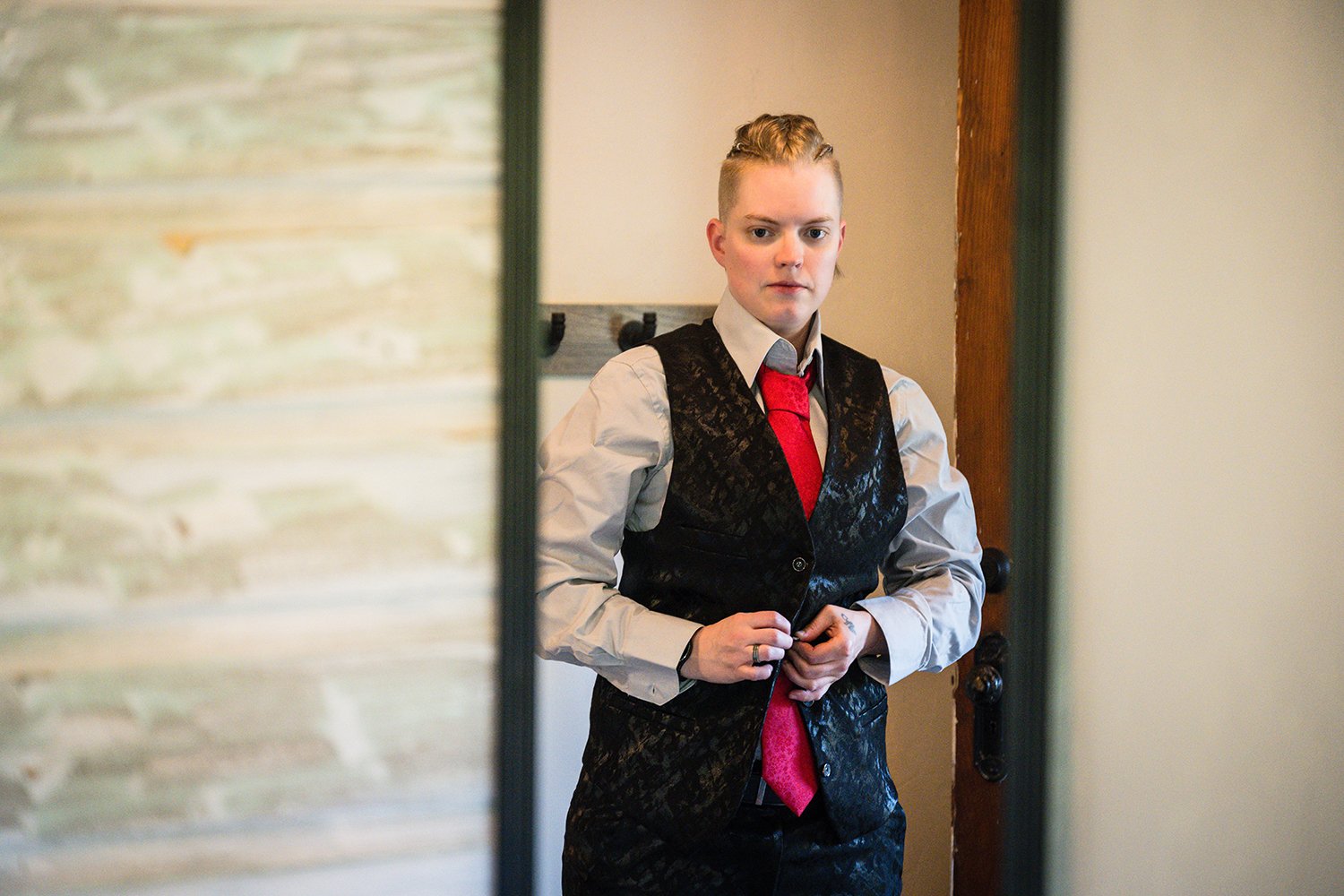 An LGBTQ+ marrier stands in front of a mirror while buttoning their floral black wedding vest and adjusting their floral red tie on her elopement day.