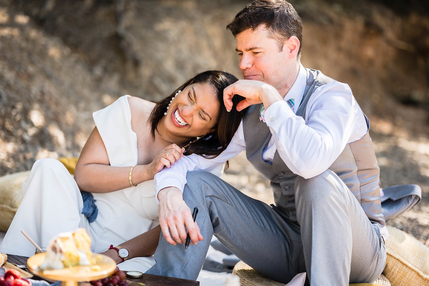 A bride laughs hard as her groom makes a funny face after eating something during their picnic that had a unpleasant taste.