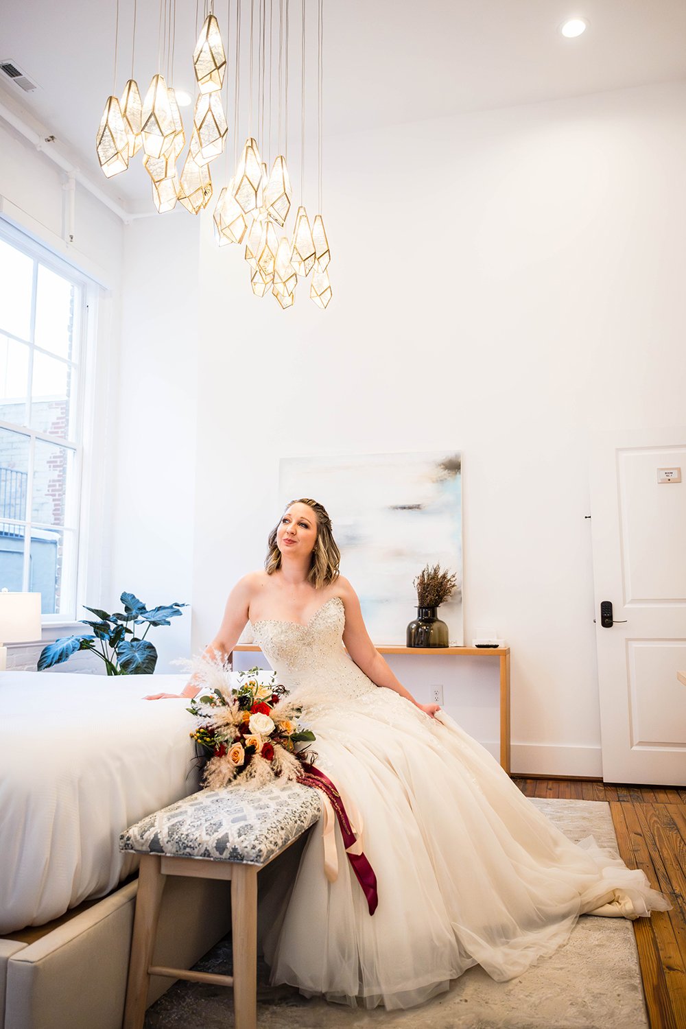 A woman in a wedding dress sits on a bench at the end of the bed in her hotel room with her bouquet by her side and looks up towards the intricate light fixtures in her room.