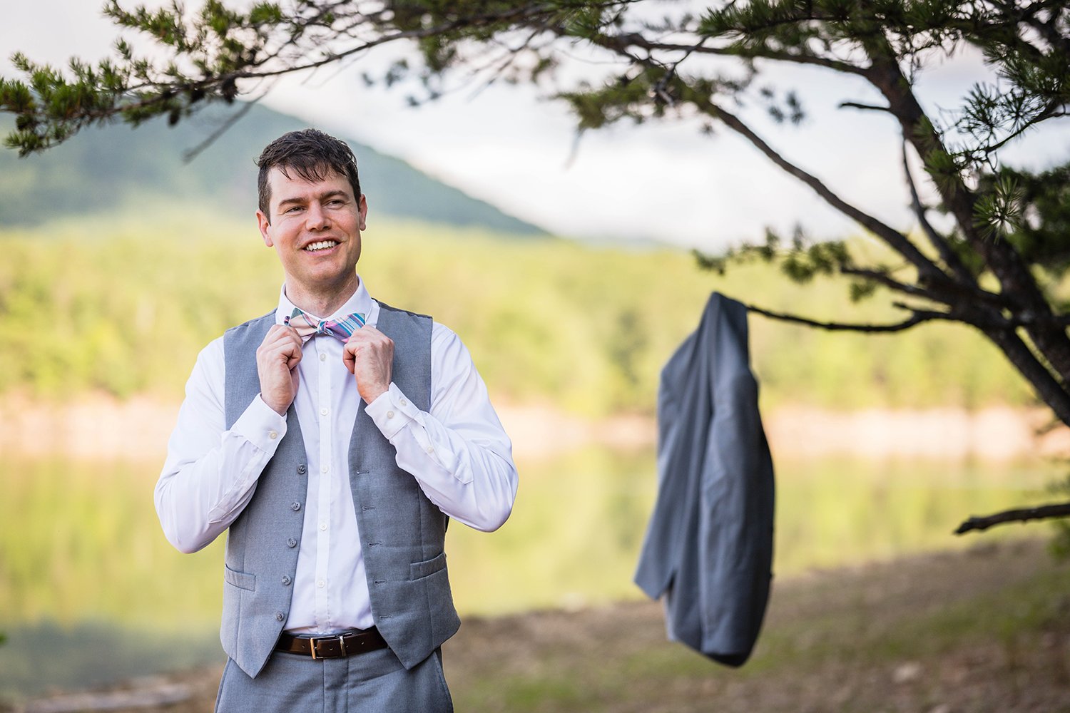A groom adjusts his bowtie and smiles looking off in the distance while his suit jacket briefly hangs on a tree branch in the background.