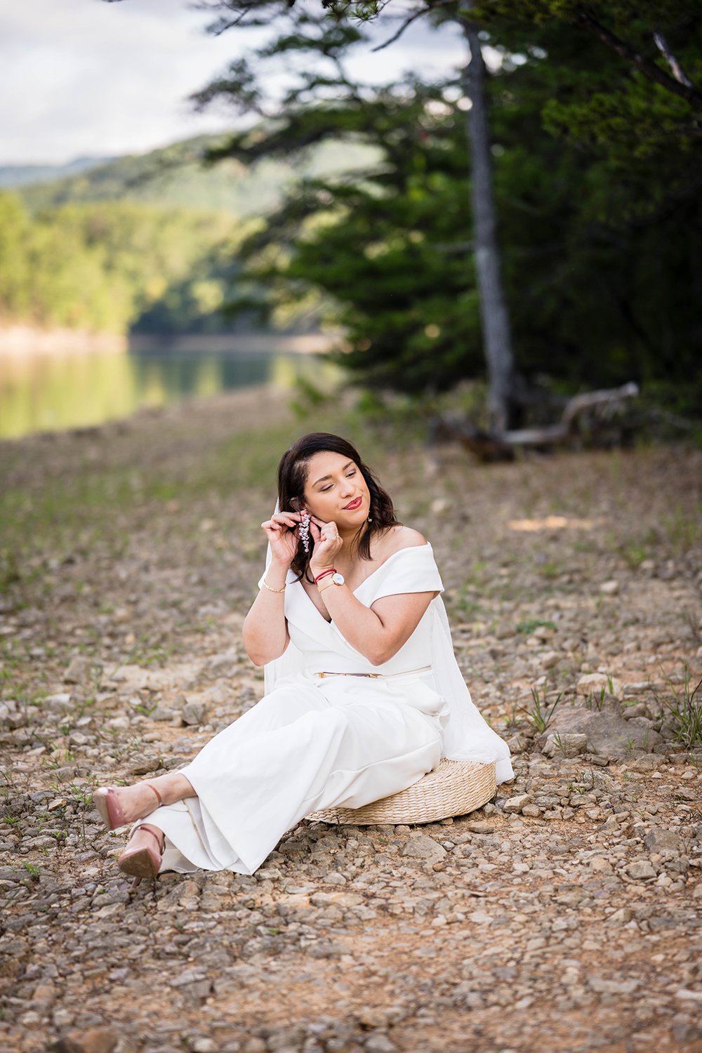 A bride on her elopement day sits on a round and close-to-the-ground wicker seat and adjusts her earring at Carvin's Cove, a location near the Blue Ridge Parkay.