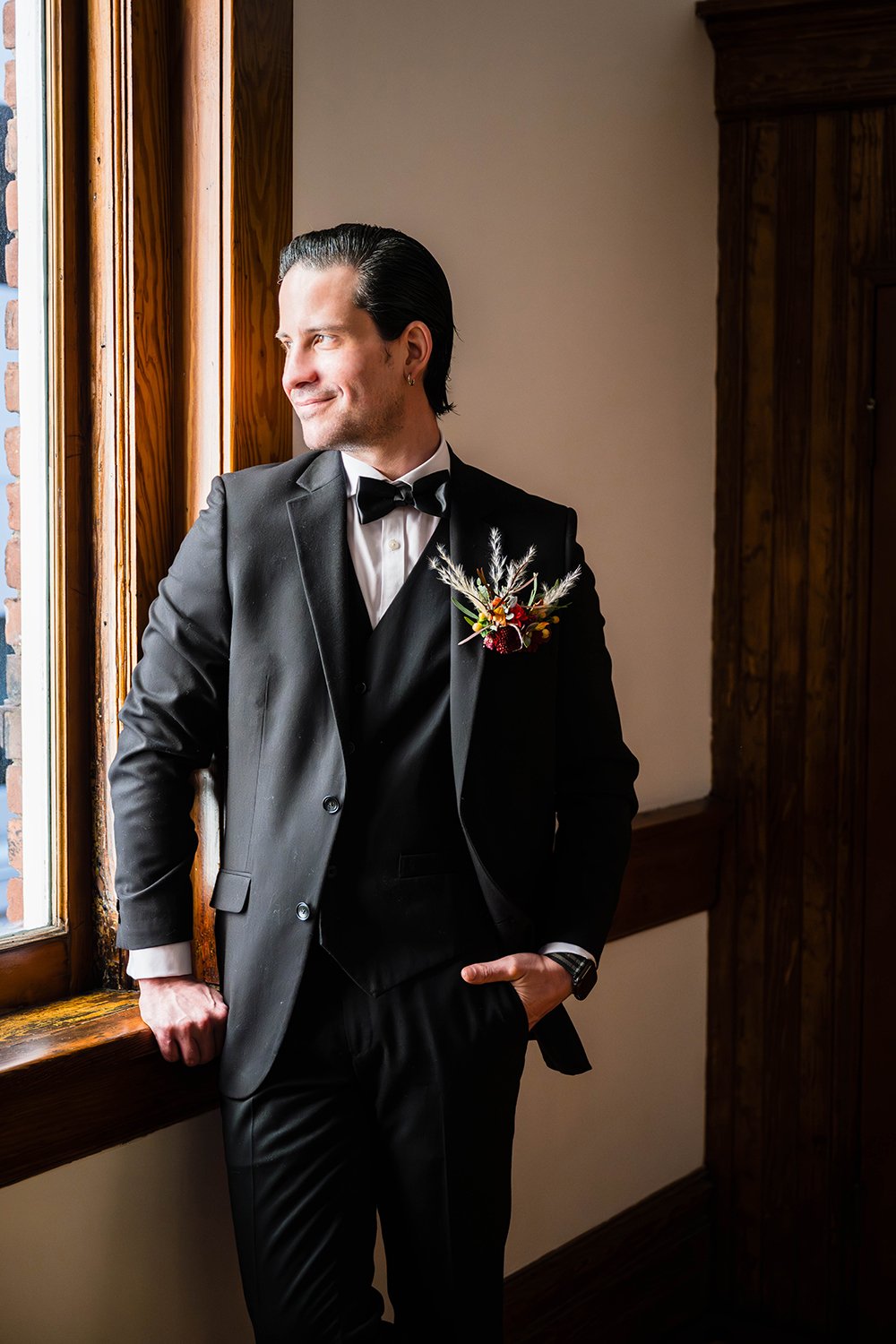 A groom stands against a wall with natural light pouring in from a window in the Bunk Room at Fire Station One for his portraits on his elopement day.