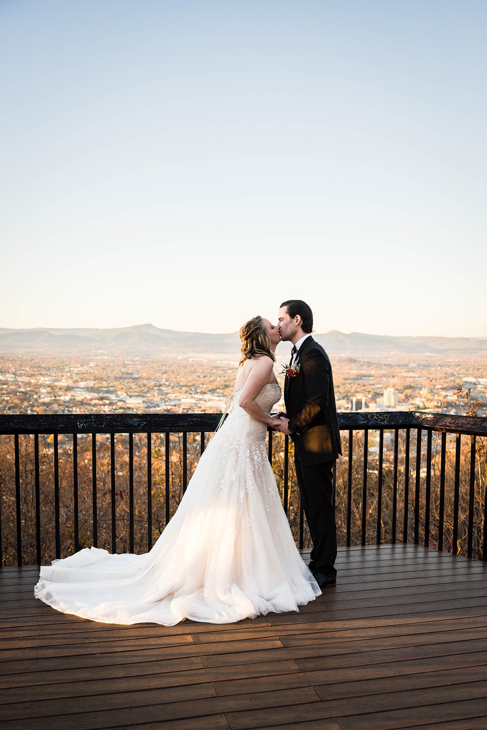 A couple on their elopement day has their first kiss at the Rockledge Overlook in Southwest Virginia, overlooking Downtown Roanoke and with the Blue Ridge Mountains vista in the background.