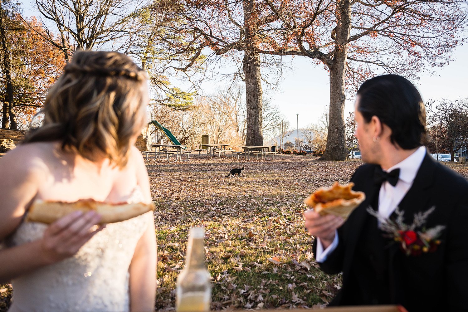 A blurred out bride and groom each holding a slice of pizza look over their shoulder to see a curious cat trying to sneak up on them while they eat at Mill Mountain Star. 