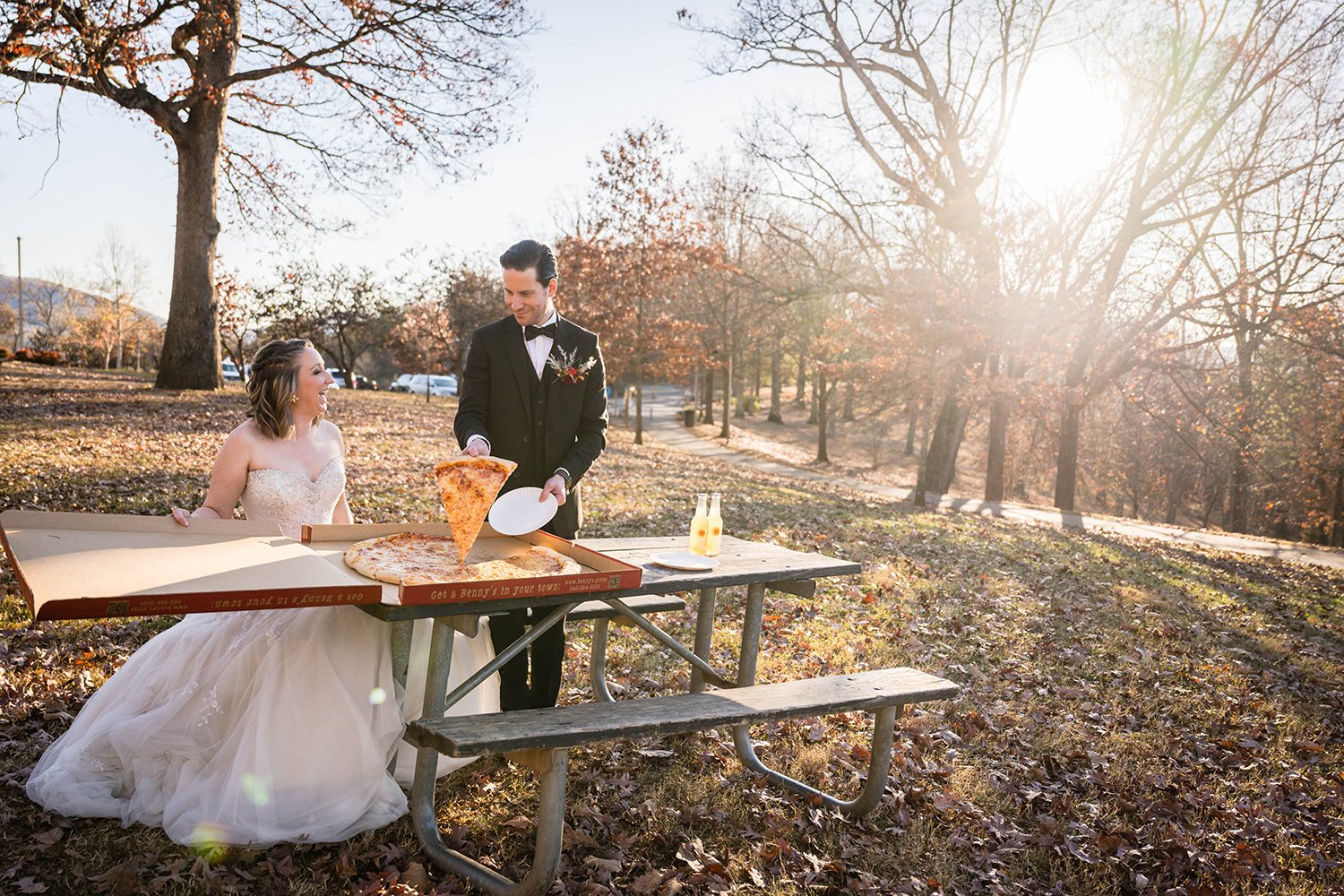  A couple places a large pizza from Benny Marconi's and two IZZE drinks on a picnic table and smile towards at one another as the groom serves a slice to his bride. 
