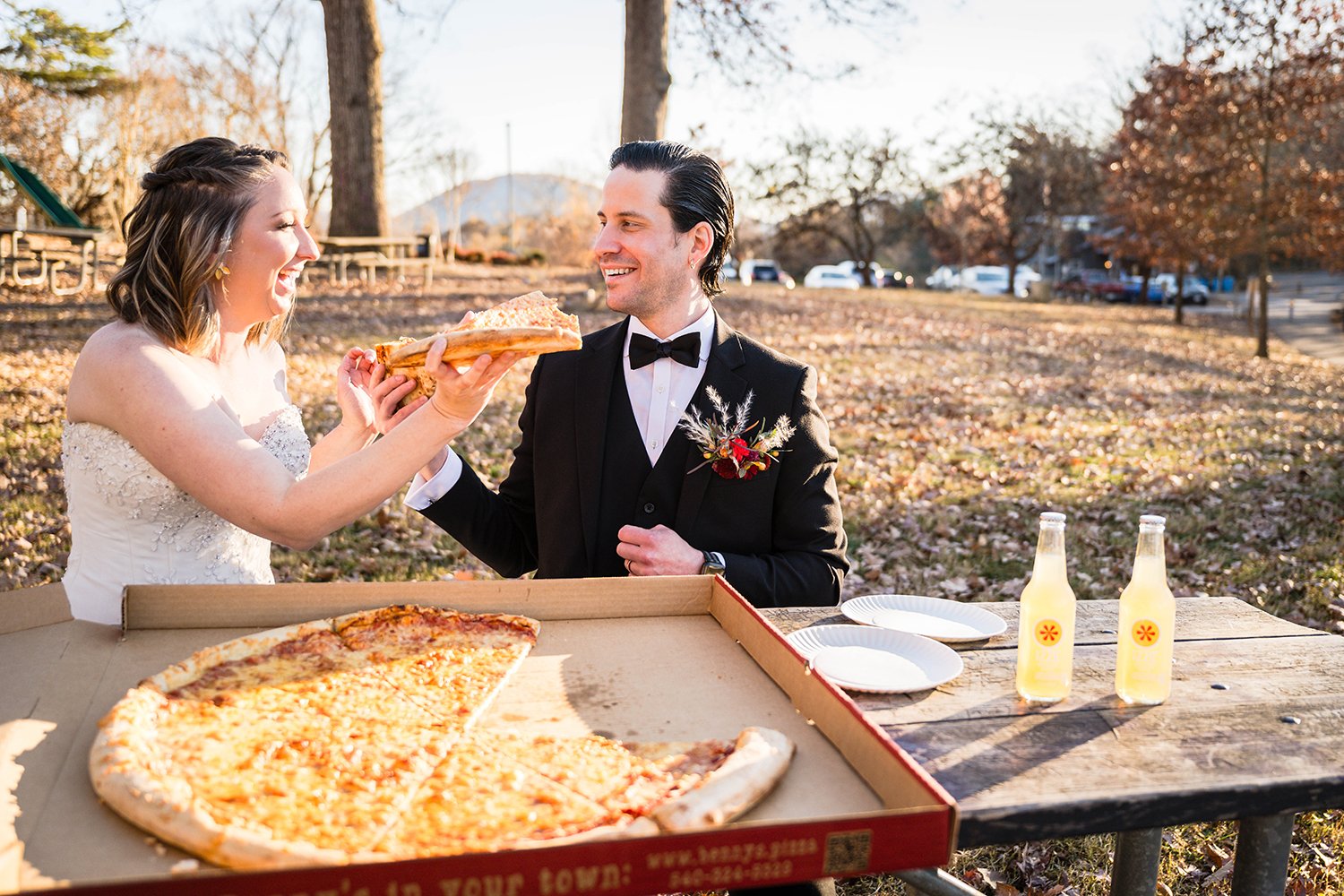 Two marriers on their elopement day at Mill Mountain Star sit at a picnic table with a Benny Marconi’s pizza and two IZZE’s and feed each other a slice.