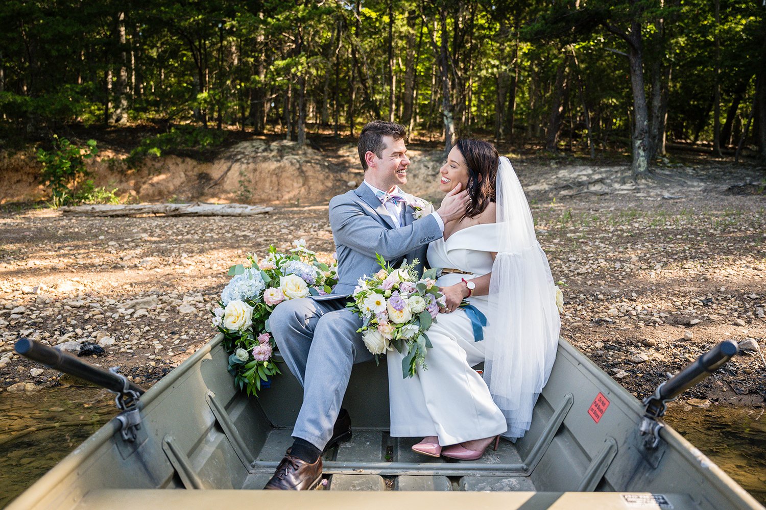 A wedding couple sits in a rowboat adorned with pastel flowers along the shoreline  of Carvin's Cove. The groom and bride smile at one another on their elopement day as the groom places his hand  on the bride's face.