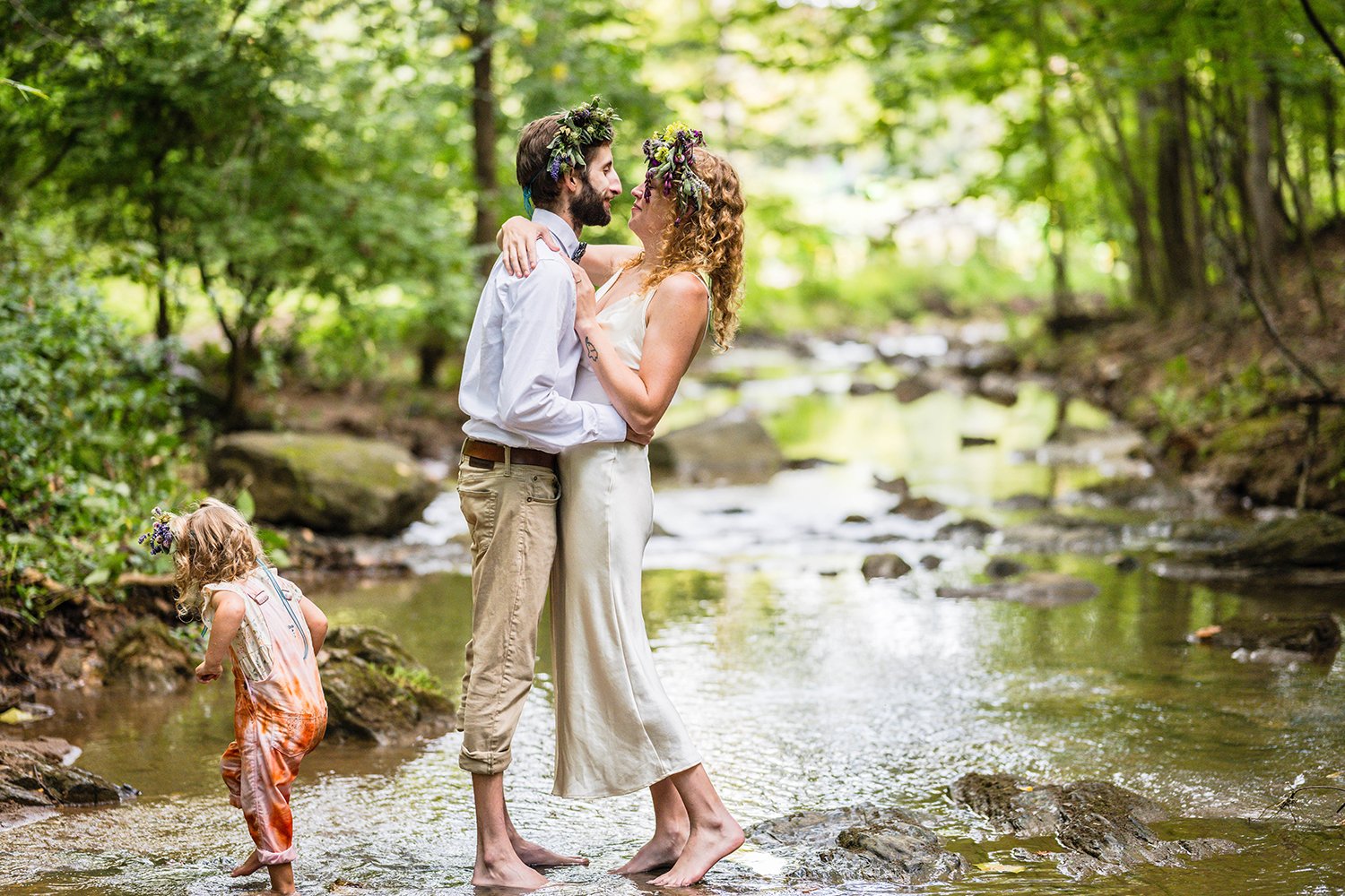 A wedding couple embrace and look lovingly into one another's arms while standing in a shallow creek under a canopy of trees as their daughter tip toes around them on their elopement day.