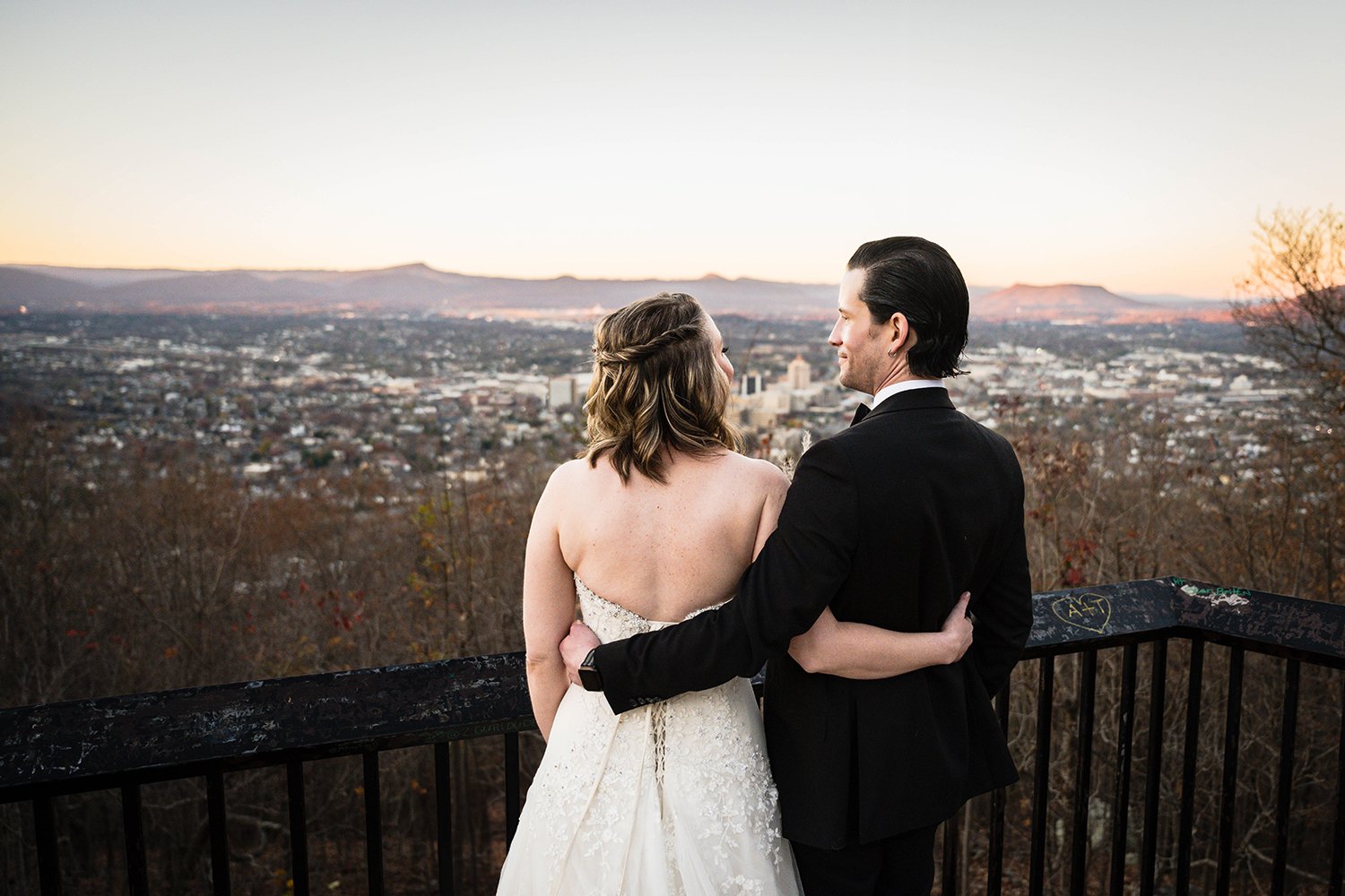 A wedding couple on their elopement day wrap their arms around one another and look at one another with the city of Roanoke and the surrounding Blue Ridge Mountains in the background.