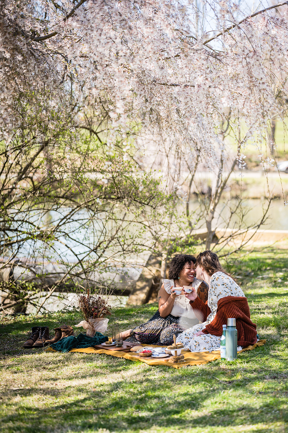 An LGBTQ+ couple snuggle up to one another during their picnic and rub noses as they clink their coffee mug under a shady tree at Duck Pond.