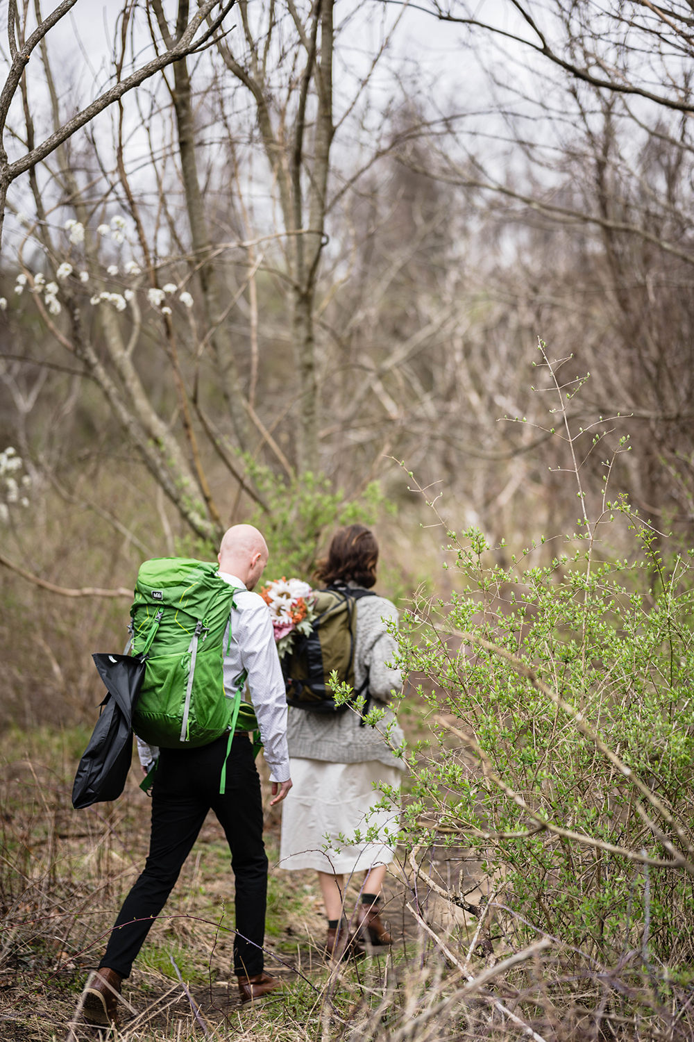 A bride wearing a hiking backpack with her bouquet leads the way on a trail followed by her groom who is also wearing a hiking backpack with a suit hanging on it.