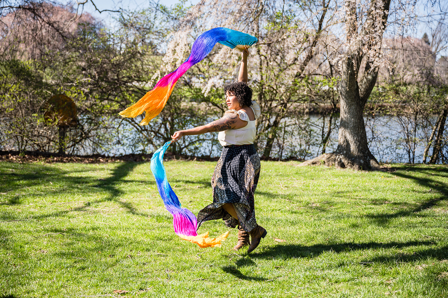 A queer BIPOC individual jumps slightly in the air as they wave their colorful silk fans during a performance at Duck Pond.