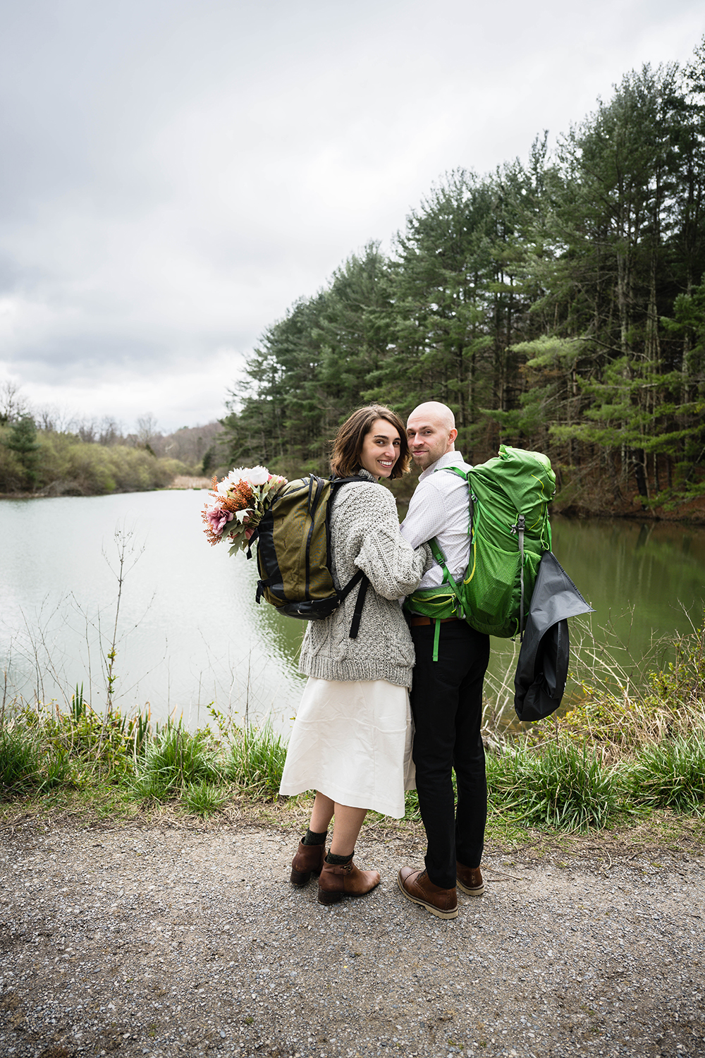 A wedding couple huddles together with their hiking gear and grabs onto one another as they look back and smile for a photo.