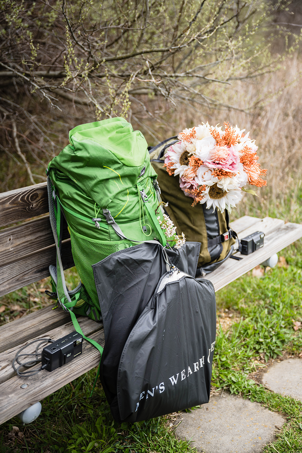 Two film cameras and two hiking backs are set on a bench at Heritage Park. One backpack is a large, green, and has a suit hanging on it while the other is slightly smaller, brown, and is holding a bouquet of fake flowers.