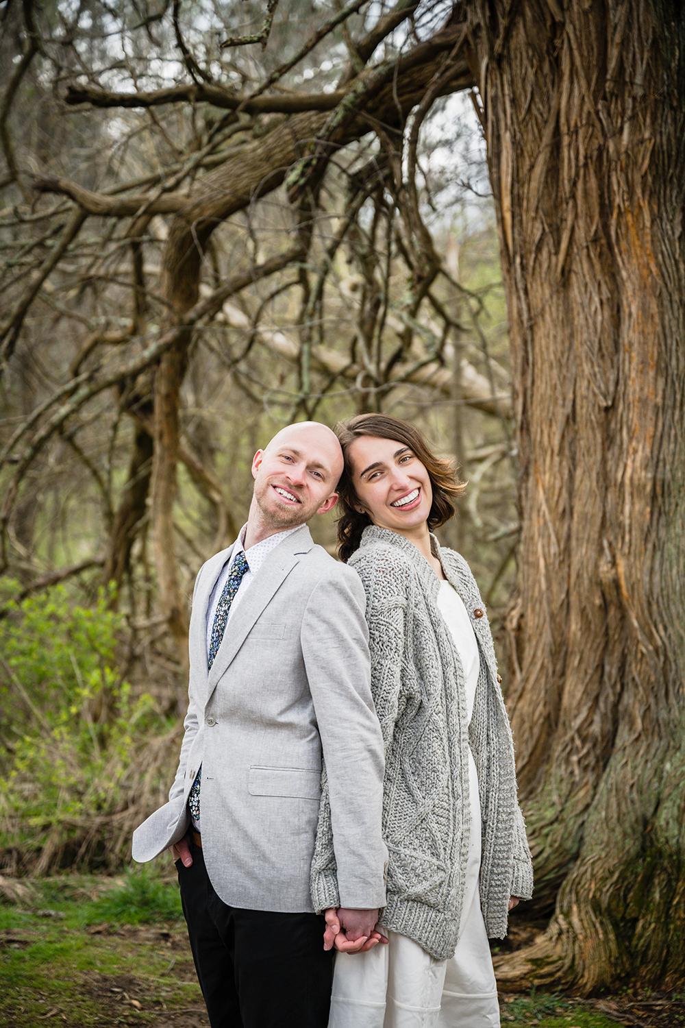 A couple hold hands in front of a tree with lots of branches and smile for a photo.