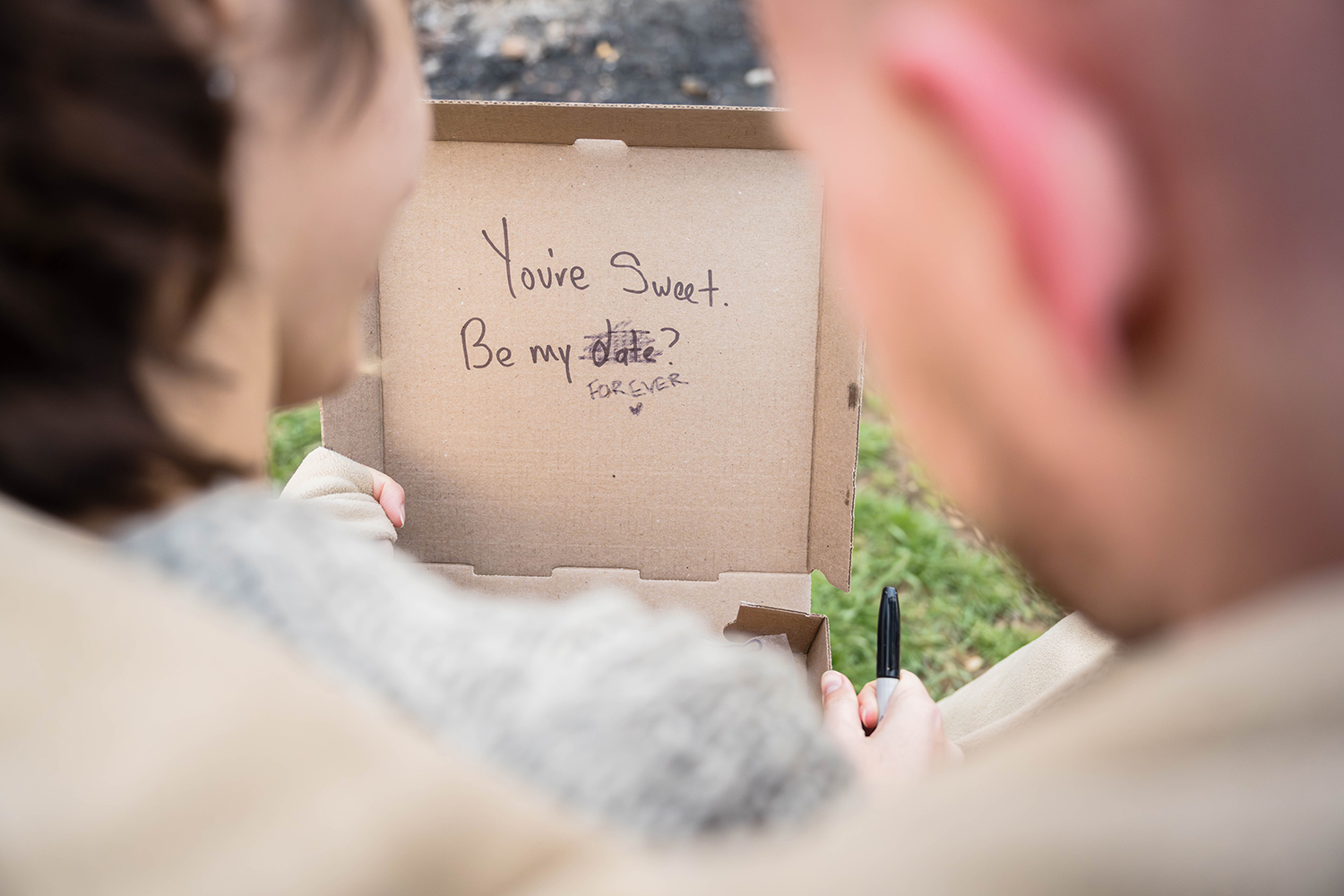 A couple cuddles together under a blanket outside of Rising Silo Brewery holding a box of cookies. On the inside of the box, someone has written "you're sweet. be my date?" with the word "date" scribbled out and replaced with the word "forever" with a little heart.