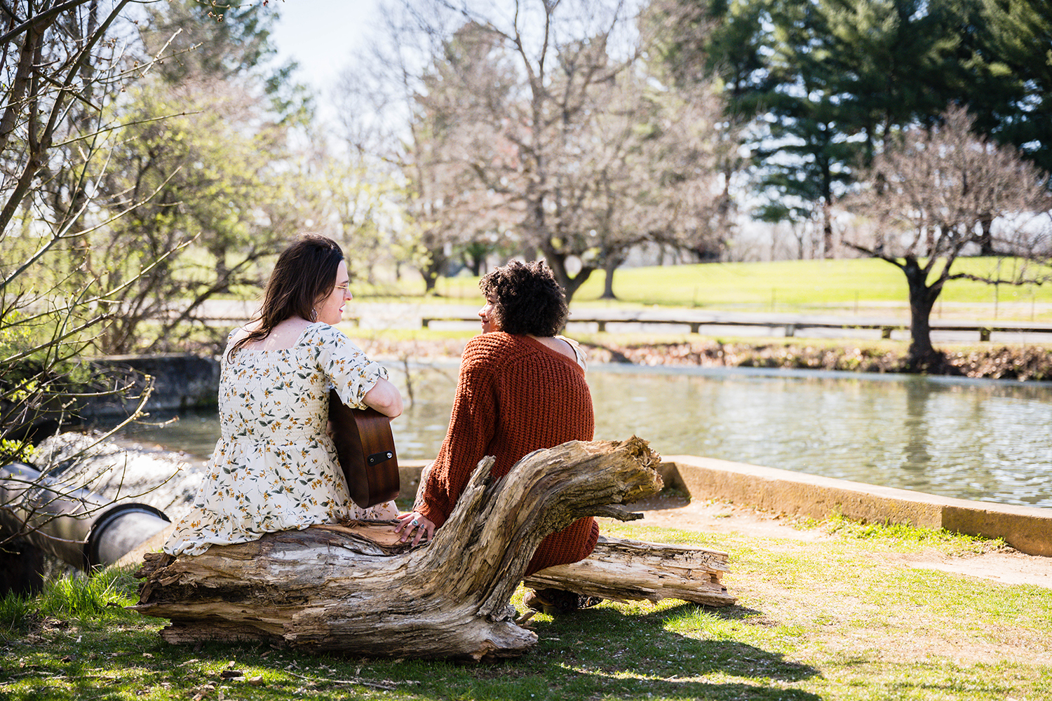 A queer couple sits on a large part of a tree at Duck Pond. One partner sings and plays the guitar while the other listens intently.