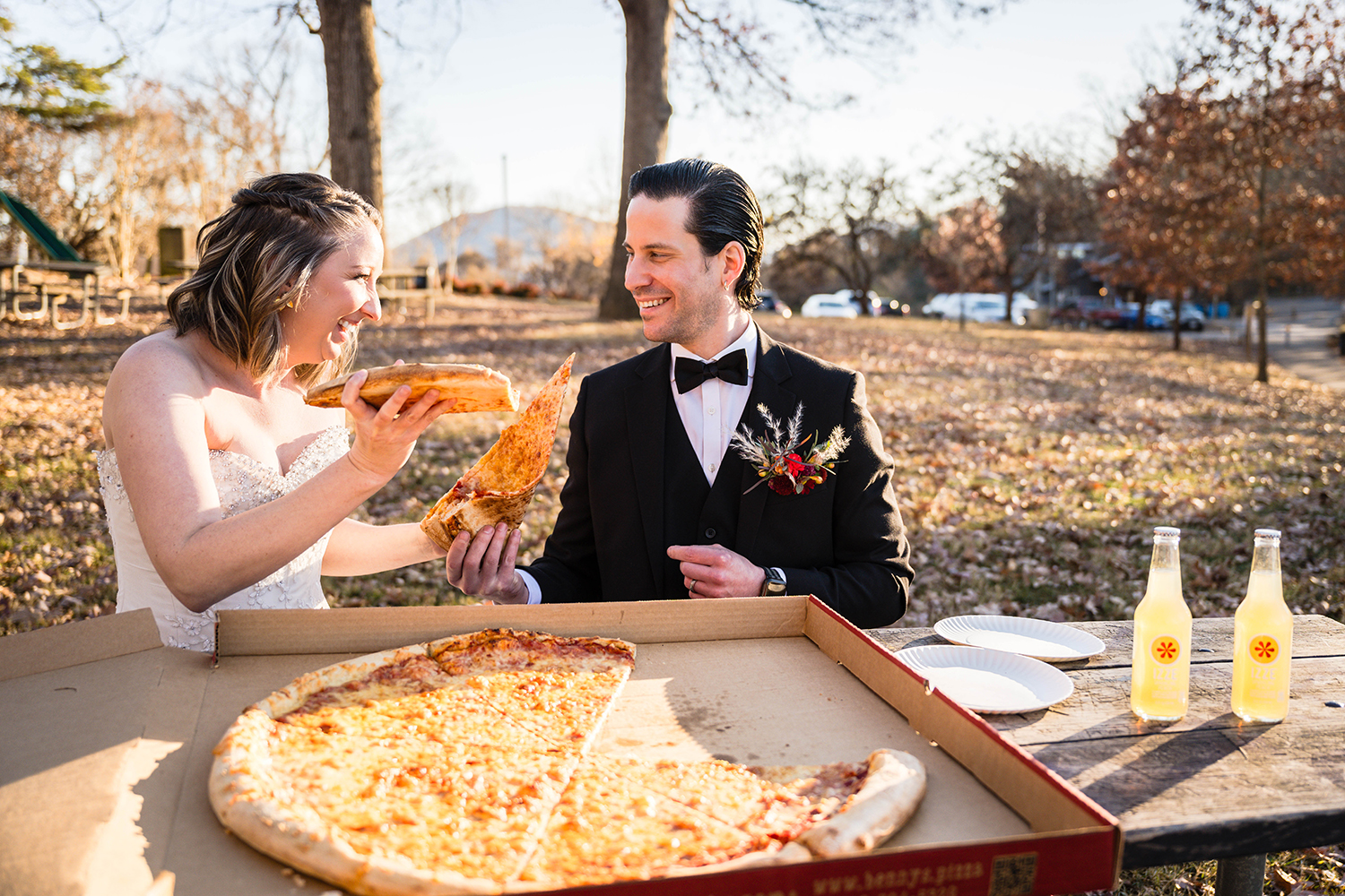 A couple on their elopement day sit at a picnic table at Mill Mountain with a box of pizza open as they each grab a slice and laugh at one another. 