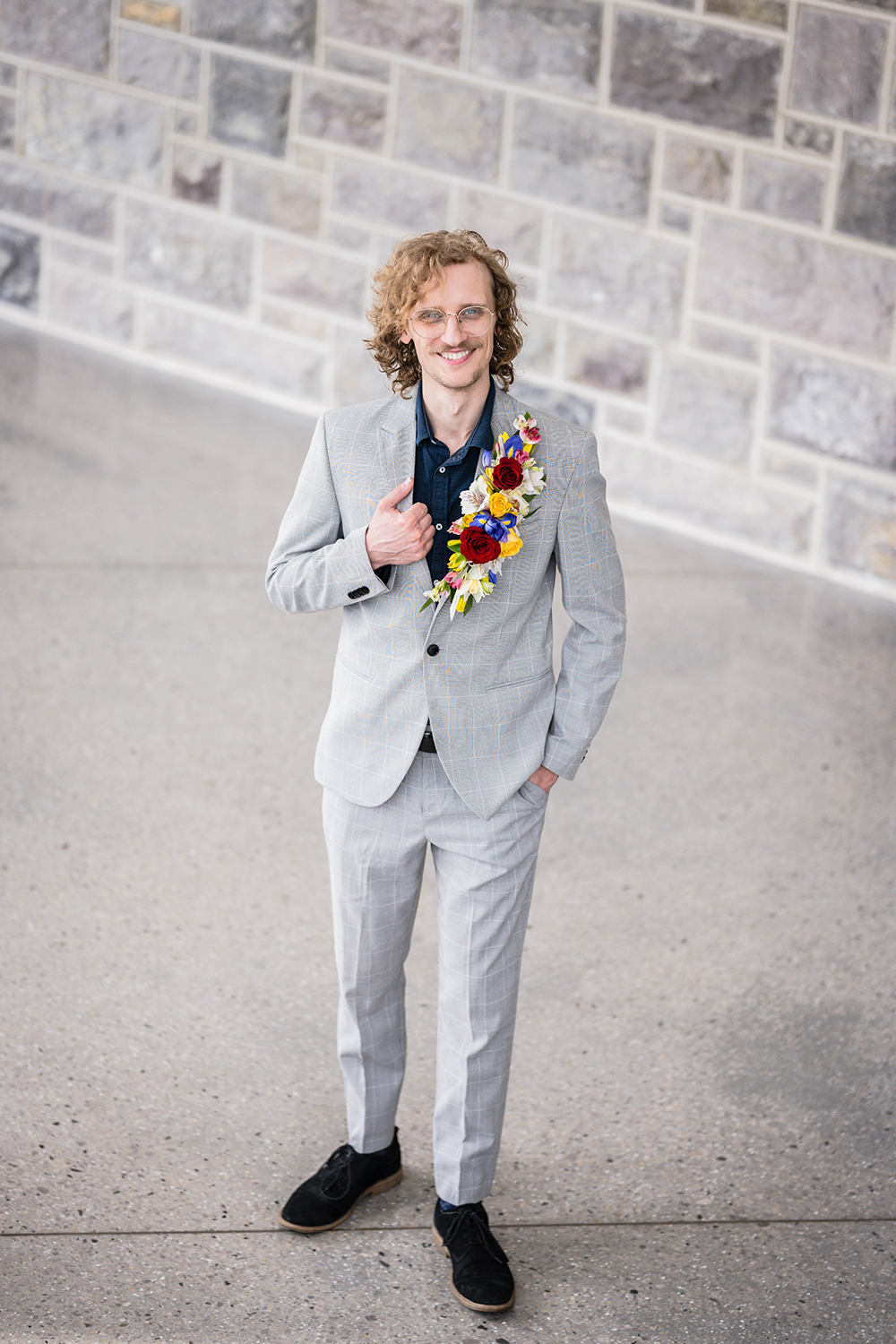 A groom wearing a gray suit with a floral lapel and navy shirt poses and smiles at the beginning of a stairwell inside of the Moss Arts Center.