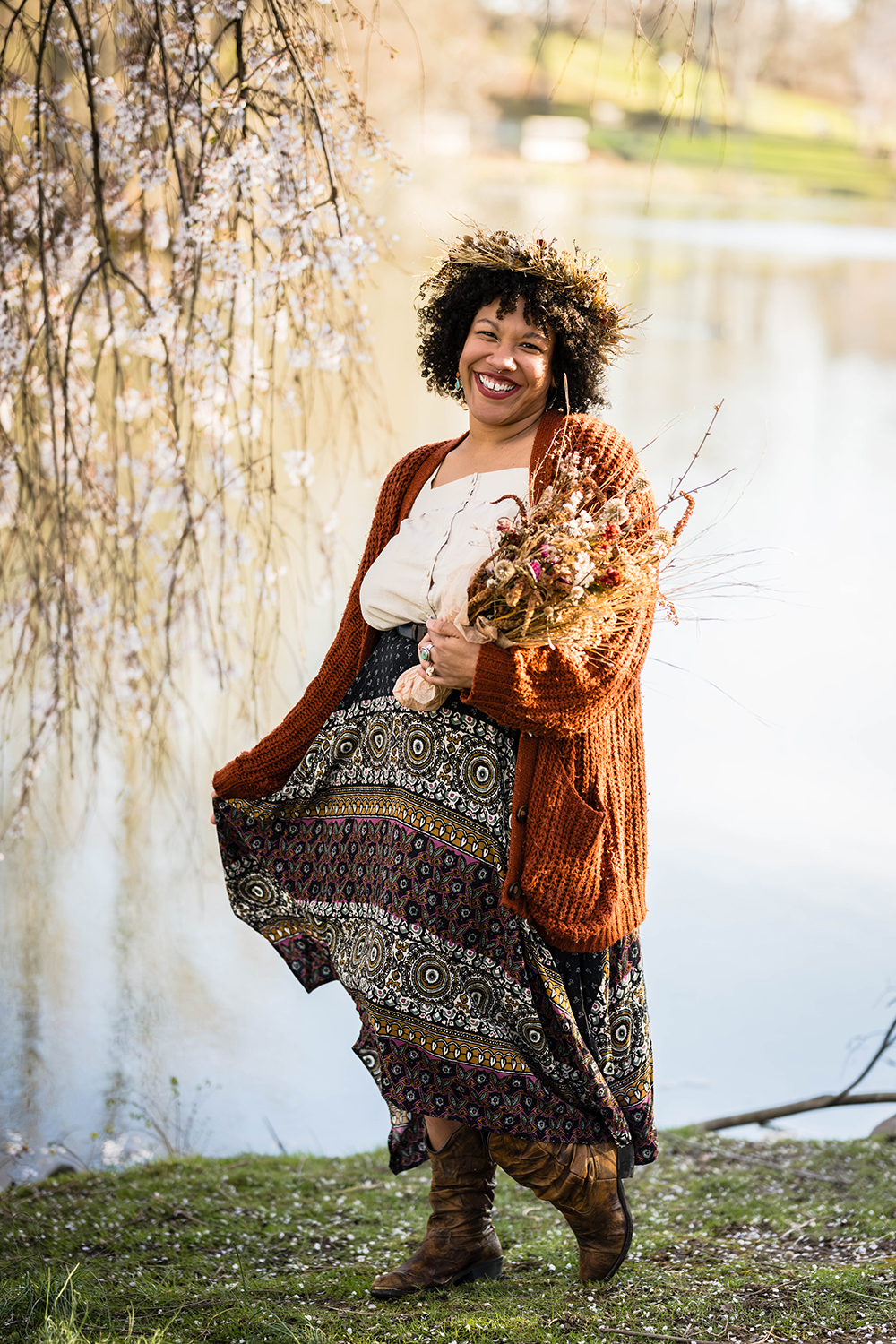 An LGBTQ+ BIPOC person wearing a dried flower crown holds a dried flower bouquet as they pose for a photo in front of a willow tree.