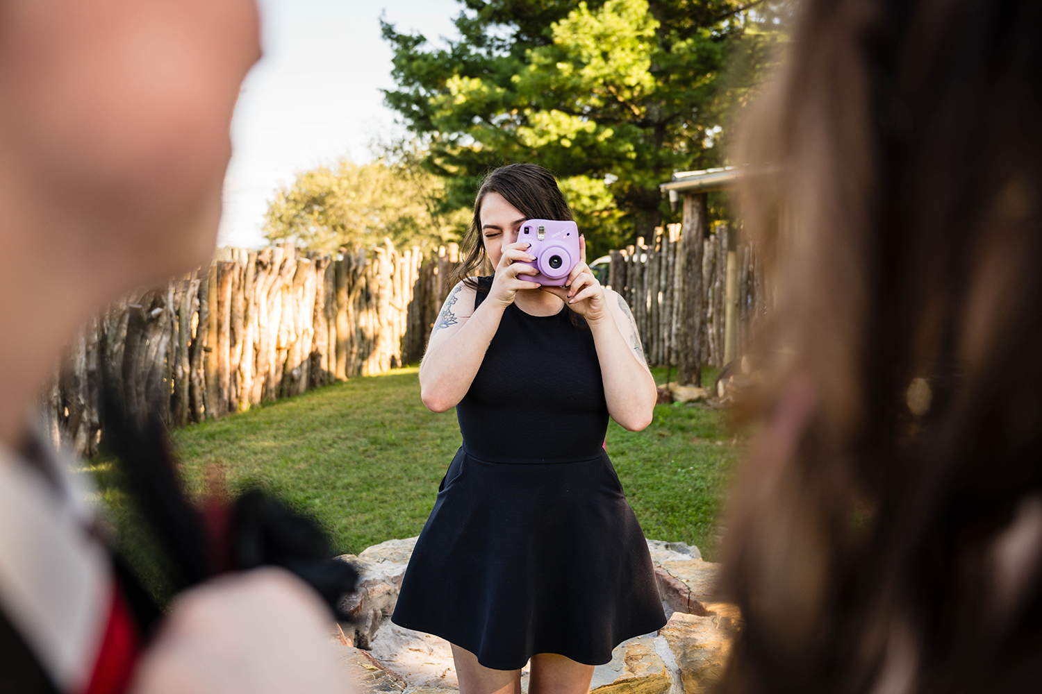 A couple blurred out in the foreground turn towards their friend holding a Polaroid camera who is ready to take a photo of the brides.