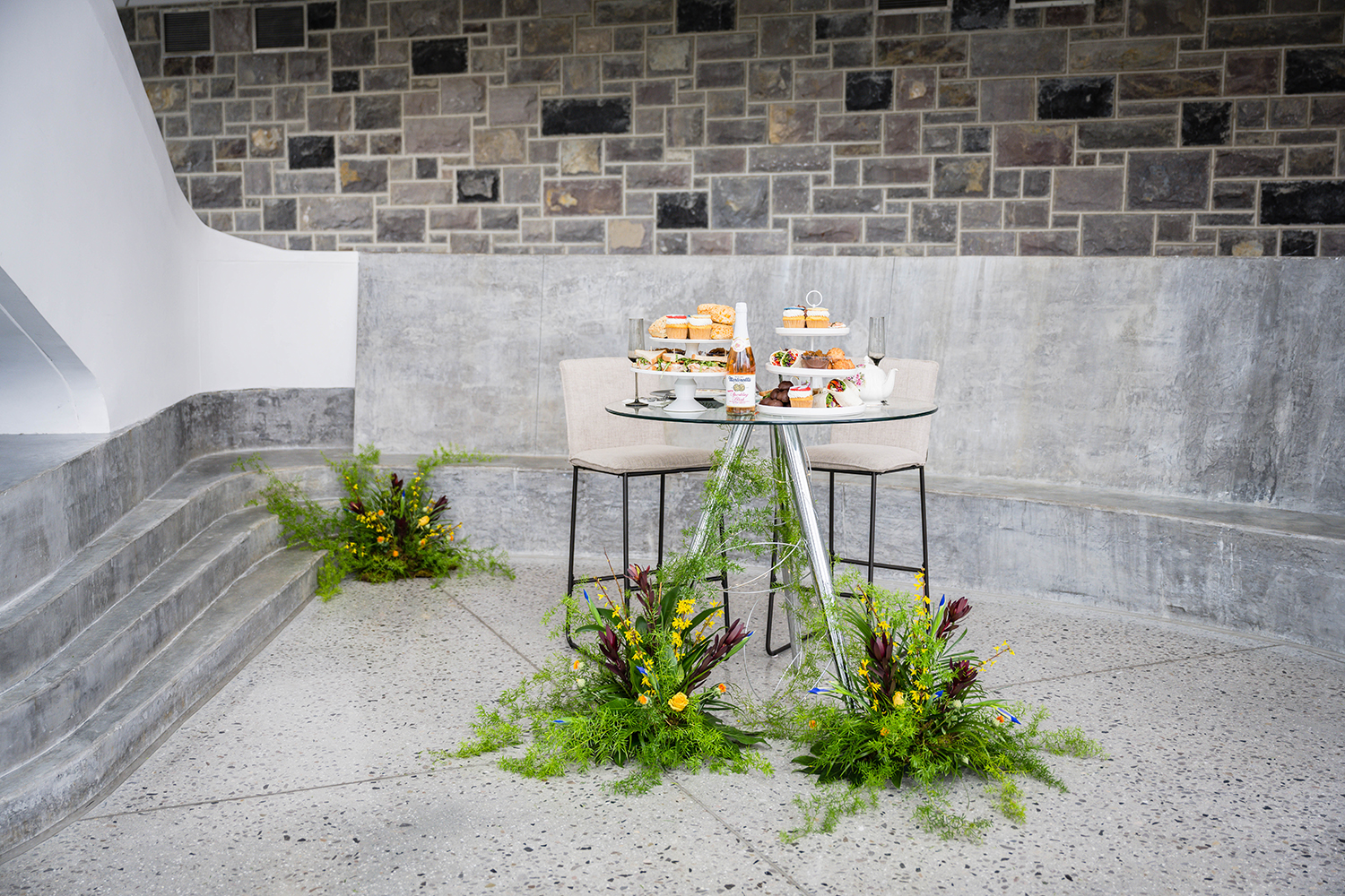A tall, clear, glass table features two tea trays full of Star-Trek inspired food and a tea set, with florals placed under the table near the legs. Two tall chairs sit empty behind the table. Another floral arrangement is placed behind the table against a wall.