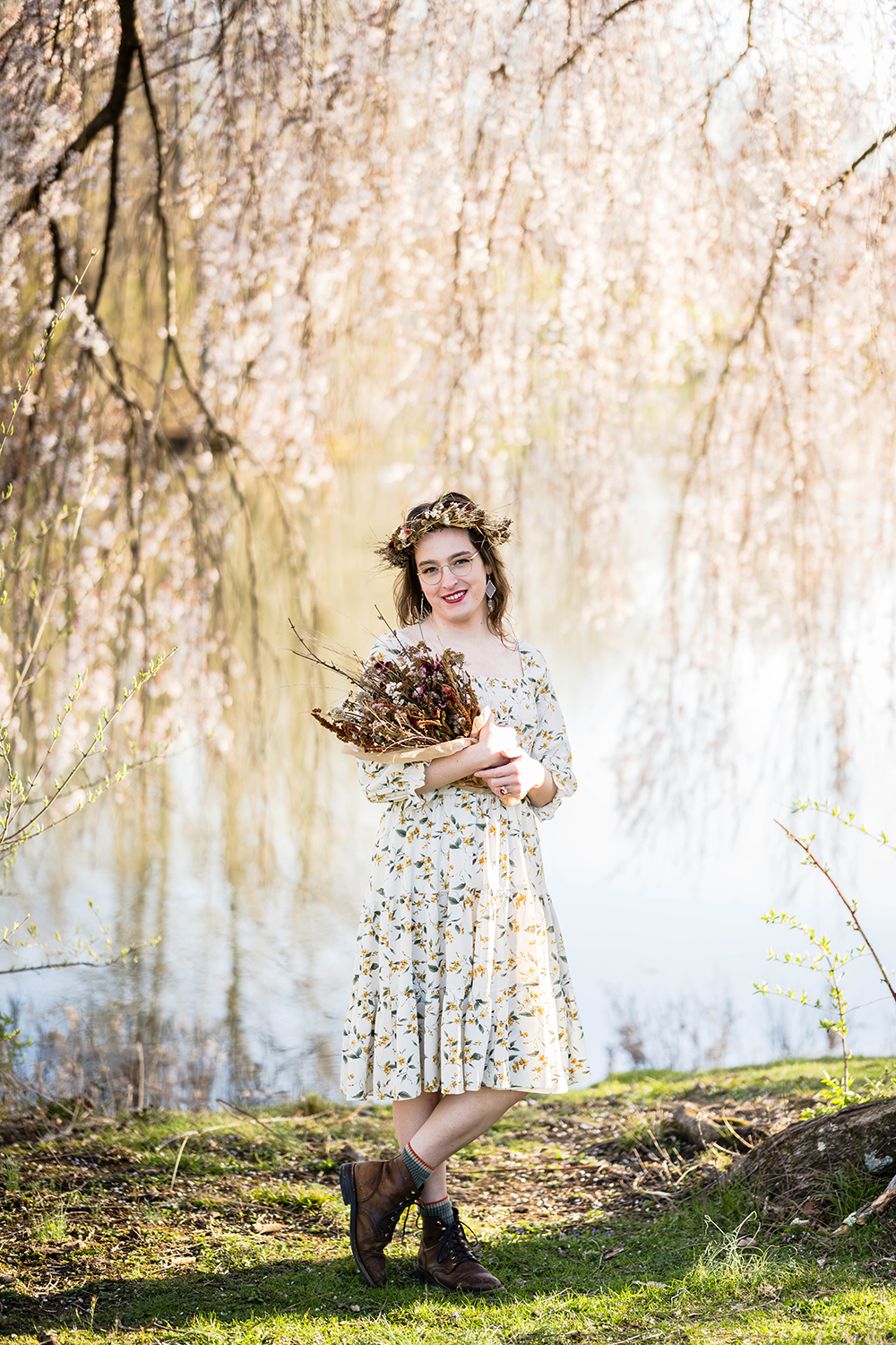 A transwoman wearing a dried flower crown holds a dried flower bouquet as she poses for a photo in front of a willow tree.