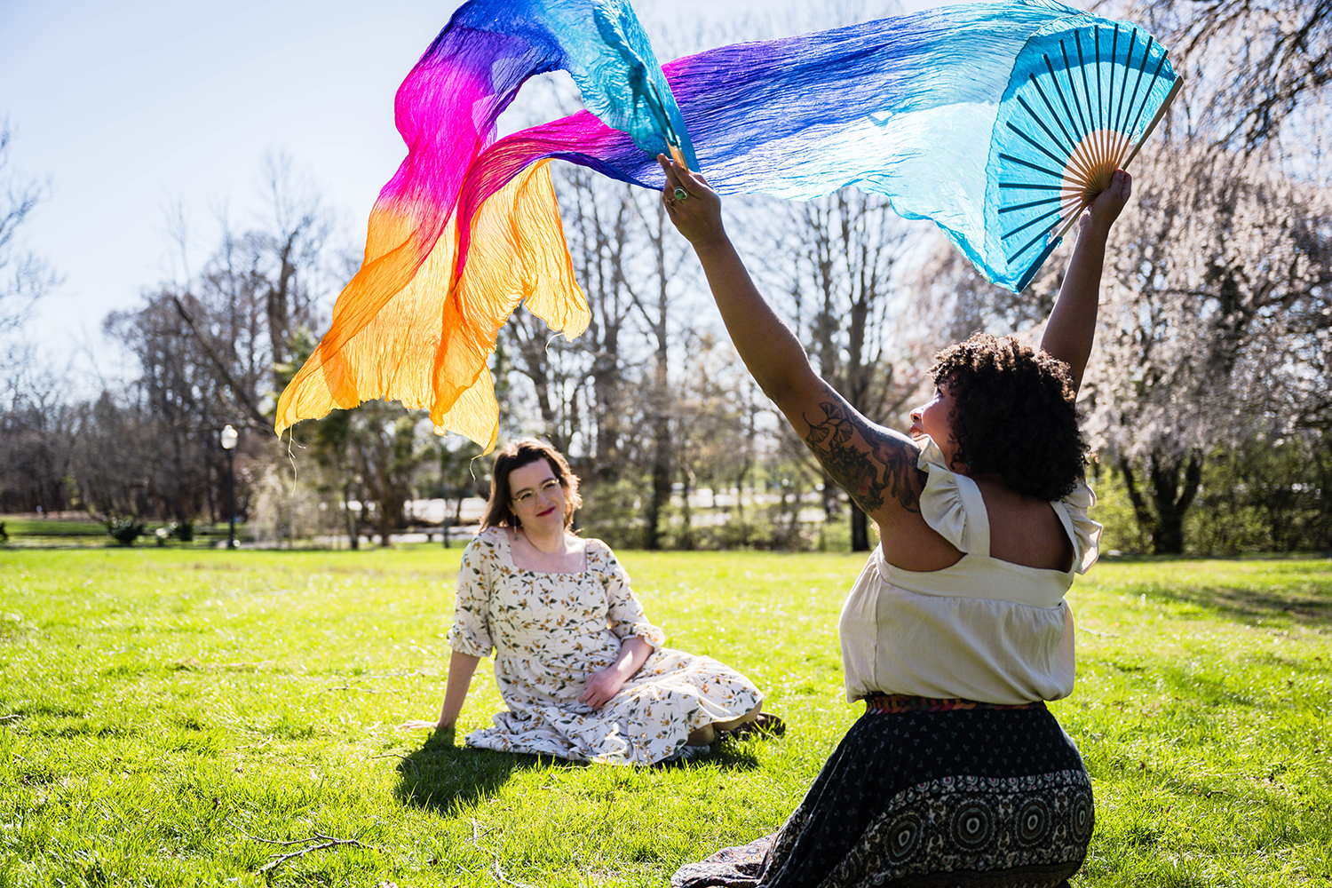 A woman sits in a field of grass and watches as their partner holds up colorful silk fans towards the sky and smiles broadly.