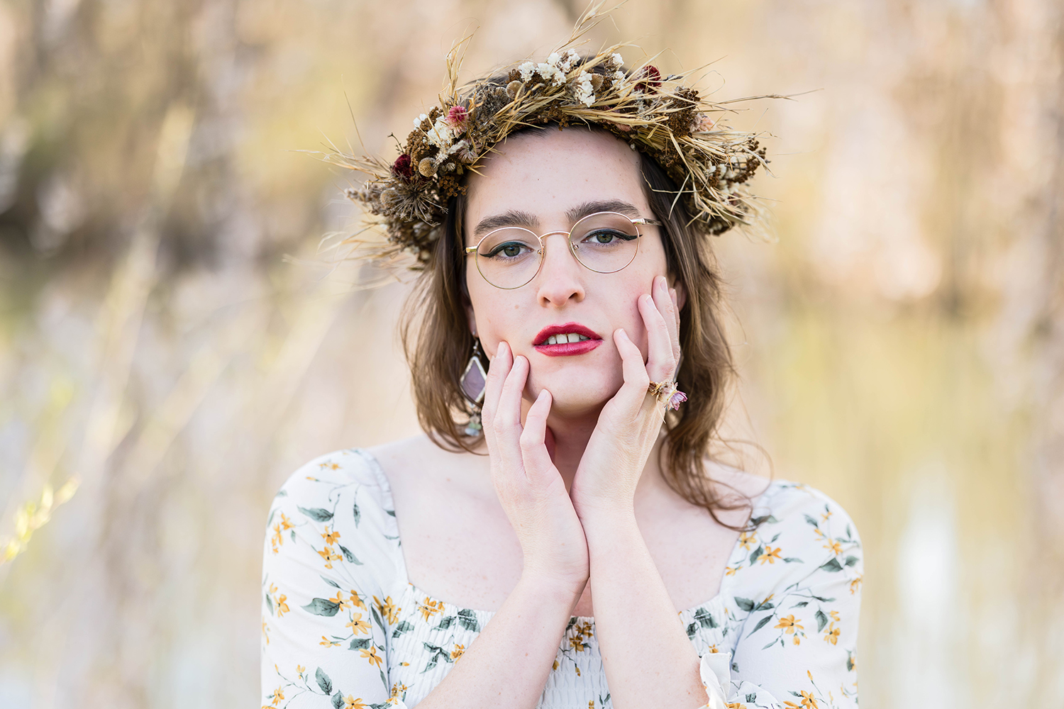 A transwoman wearing a dried flower crown touches her face as she poses for a photo in front of a willow tree.