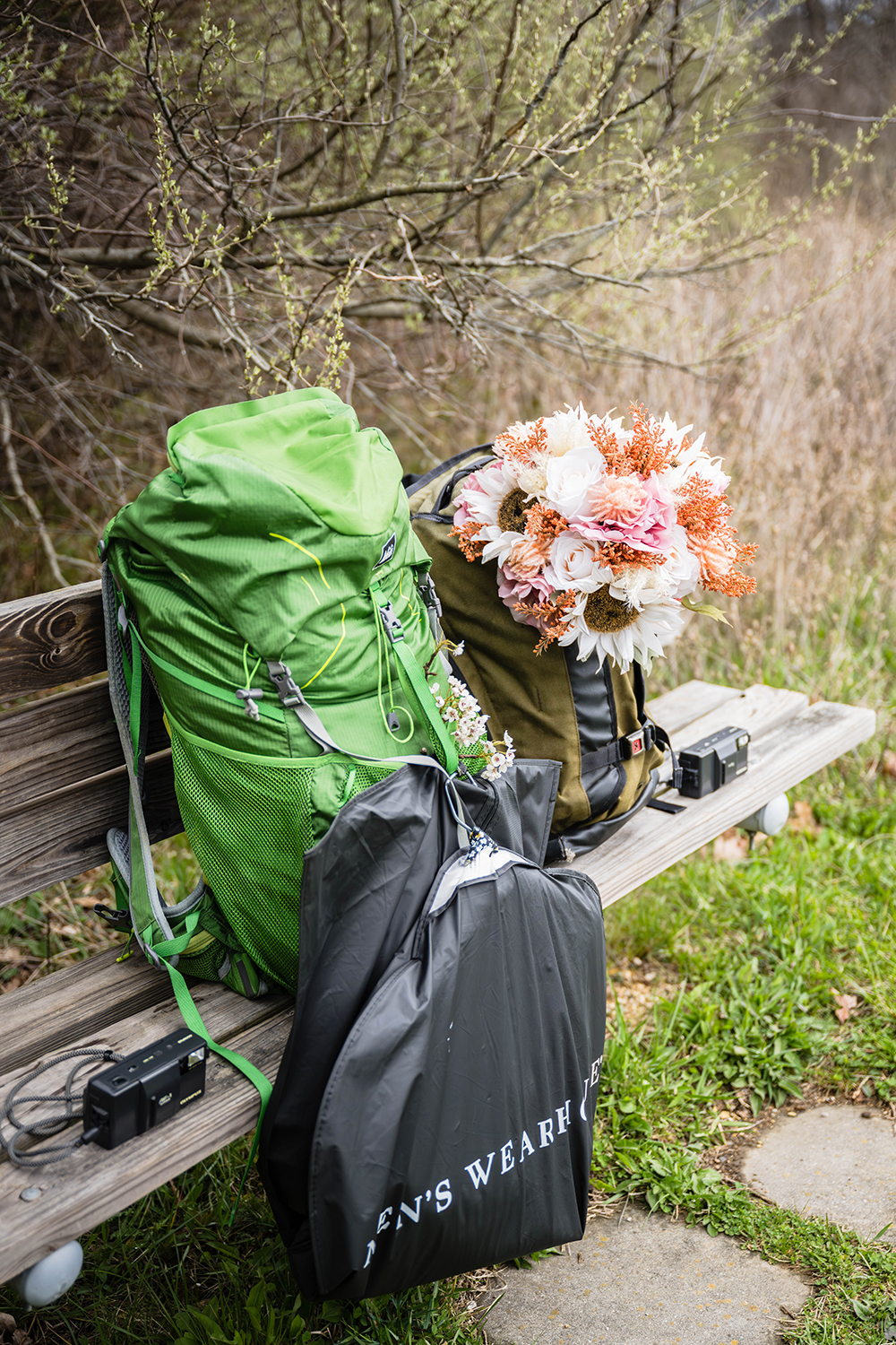 Two backpacks, one with a suit jacket on a hanger pinned to the front of the bag and the other with a wedding bouquet secured in the front pocket, are placed on a bench along with two film cameras.