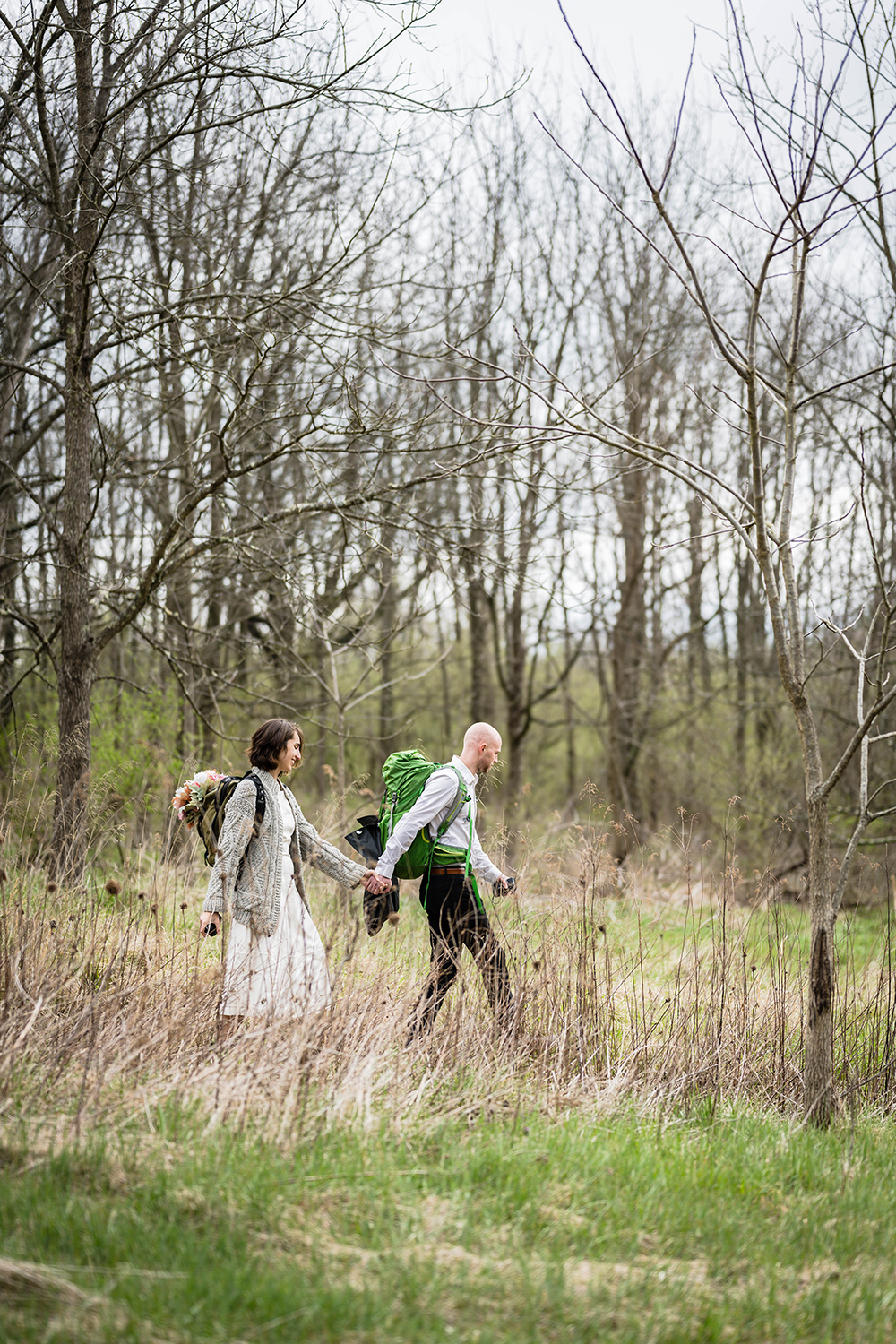 A partner wearing a backpack with a suit jacket clipped to the back holds hands with his partner and leads her through a section of the trail. The partner in the back is also wearing a backpack with a wedding bouquet strapped inside of a pocket.