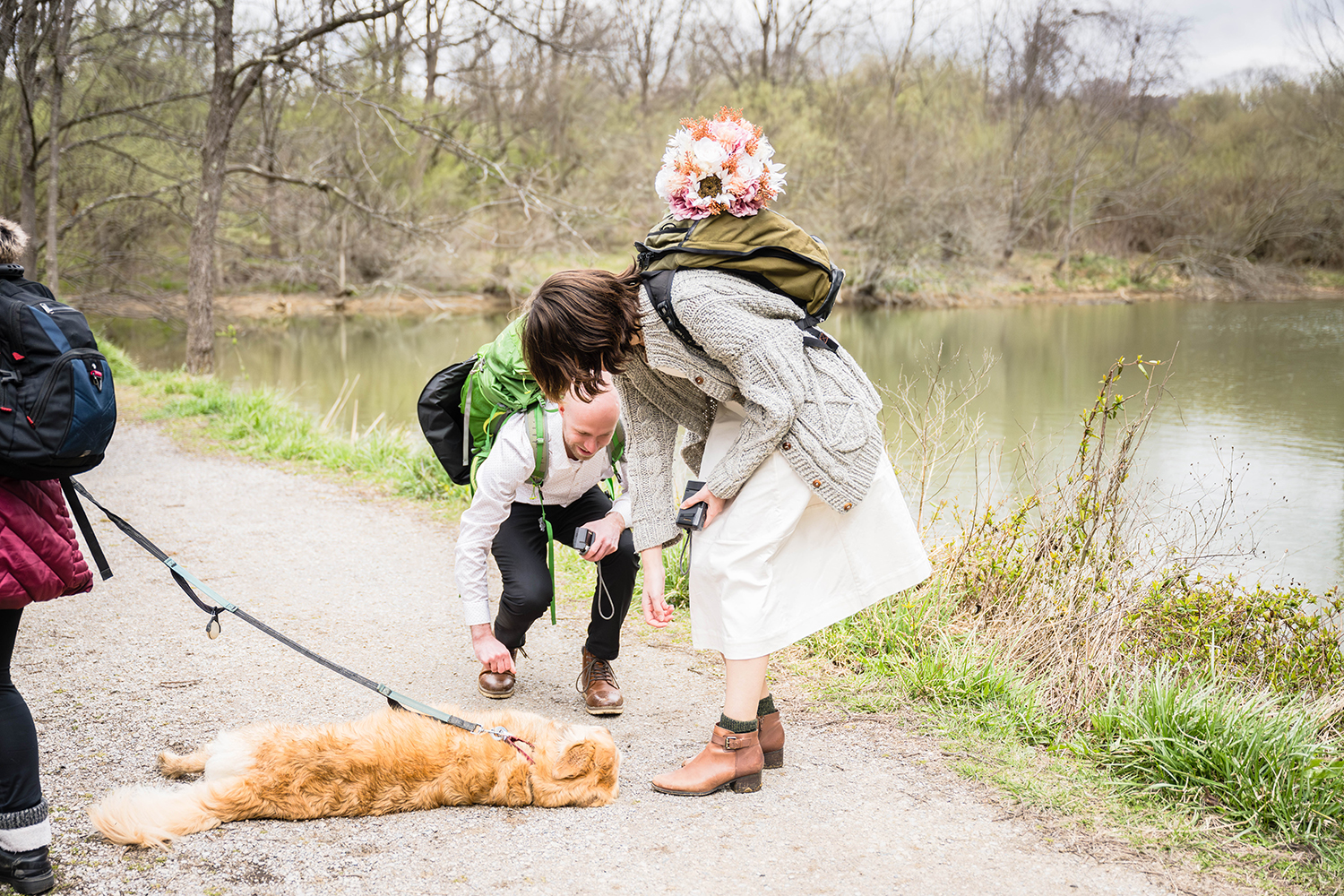 A bride and groom bend down slightly to pet a leashed dog that is lying happily on the trail.