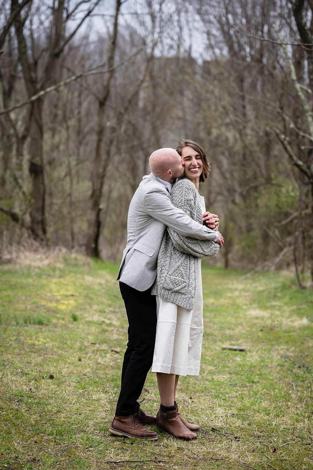 A groom hugs his bride and kisses her cheek while she smiles widely. The pair are standing in the middle of a trail on the Appalachian Trail in Virginia.