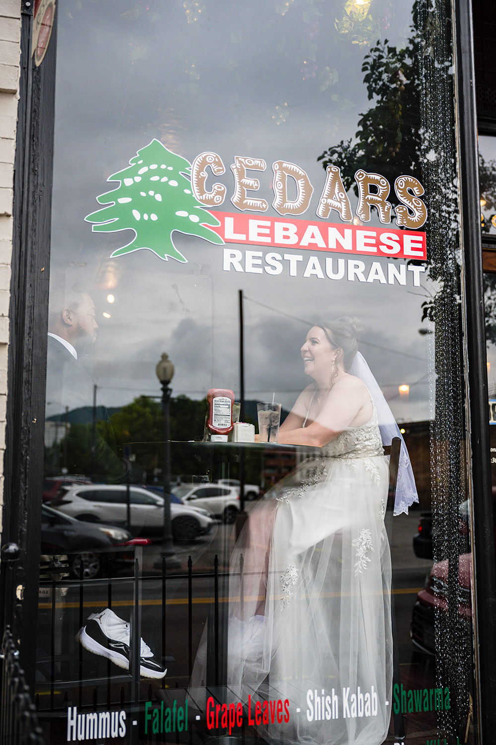 A couple wearing wedding attire sits at a high top table near a large window at Cedar's Lebanese Restaurant in Roanoke, Virginia.