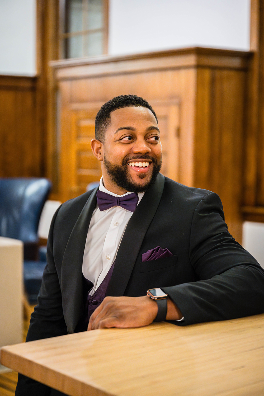 A man wearing a suit and bowtie sits at a bar inside of a hotel and smiles away from the camera.