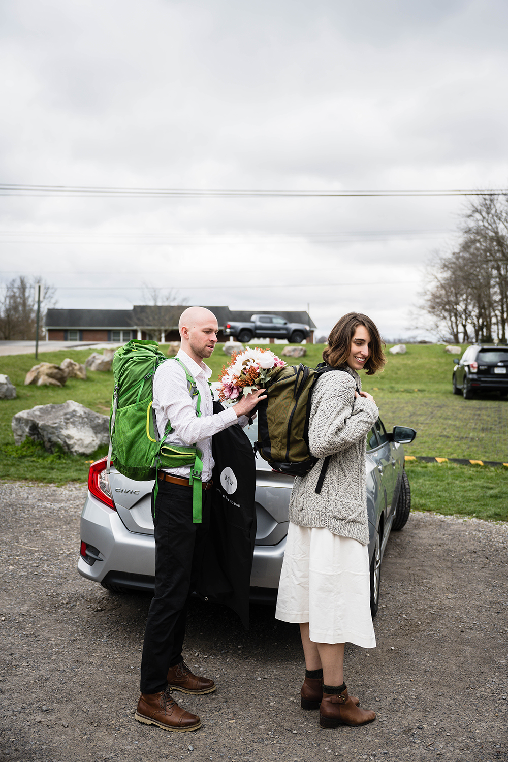 A couple on their elopement day gear up outside of their car at the trailhead parking lot. Both marriers are wearing backpacks. One partner is holding their suit jacket while helping their significant other secure her bouquet in her day bag.