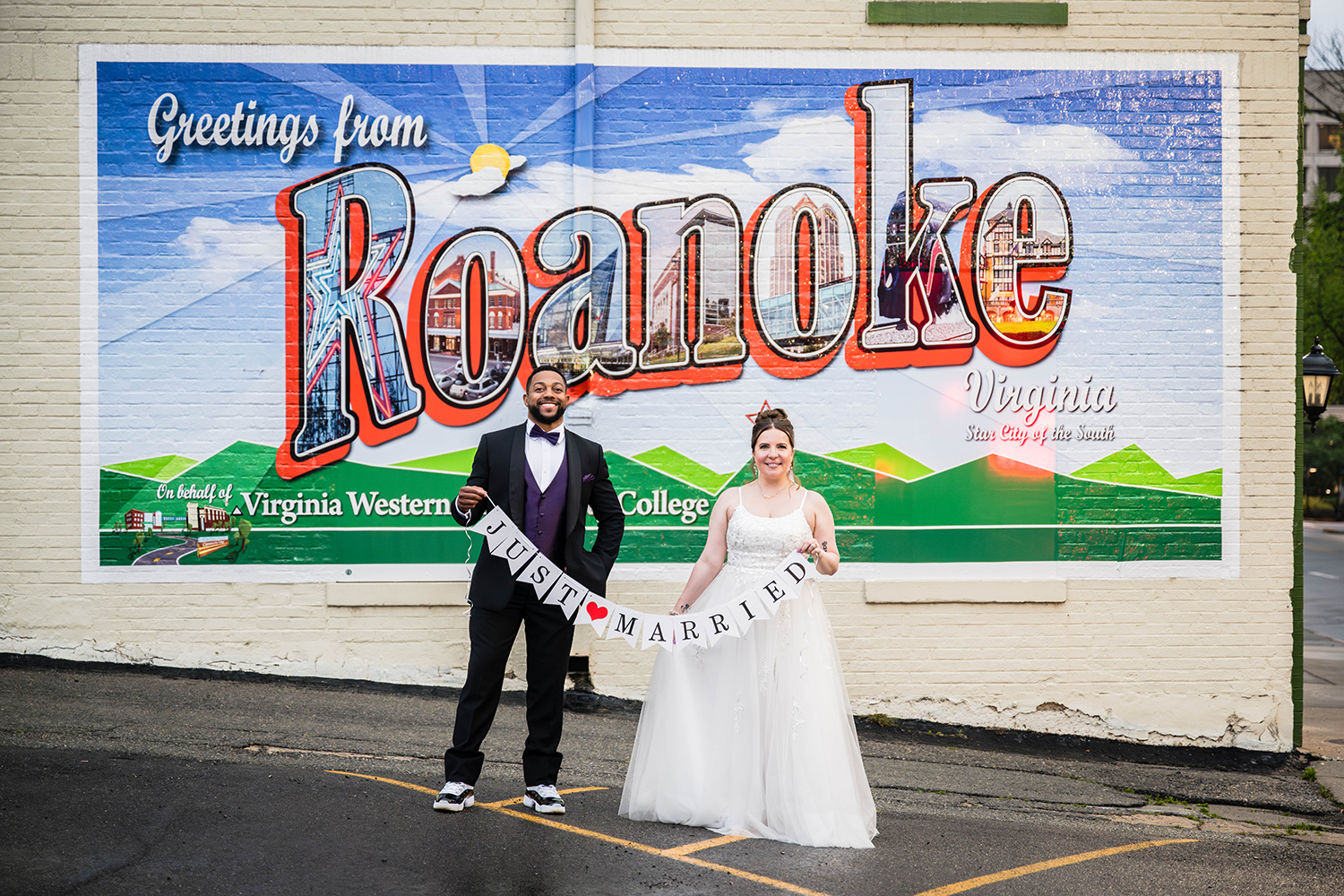 A couple wearing wedding attire stands in front of a mural that says "Greetings from Roanoke, Virginia: Star City of the South" holding a banner that reads "just married".