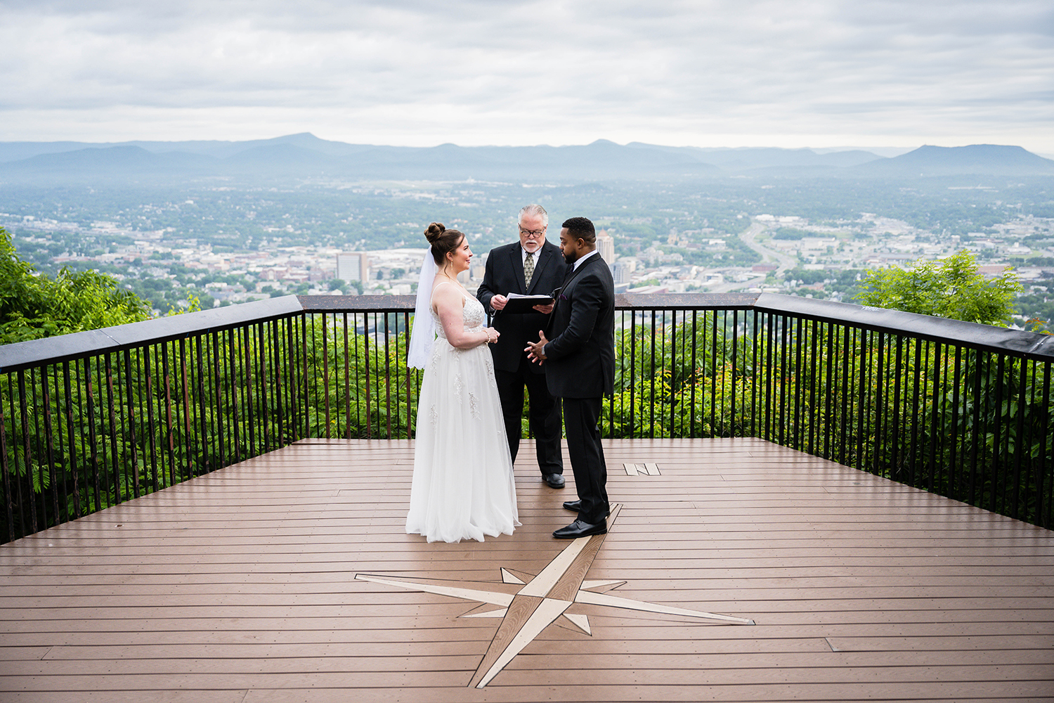 A couple on their elopement day stand in front of their officiant as their sunrise ceremony begins at Mill Mountain Park. The couple and officiant are standing at the Mill Mountain Star Overlook, which features the Roanoke city skyline and the surrounding Blue Ridge Mountains.