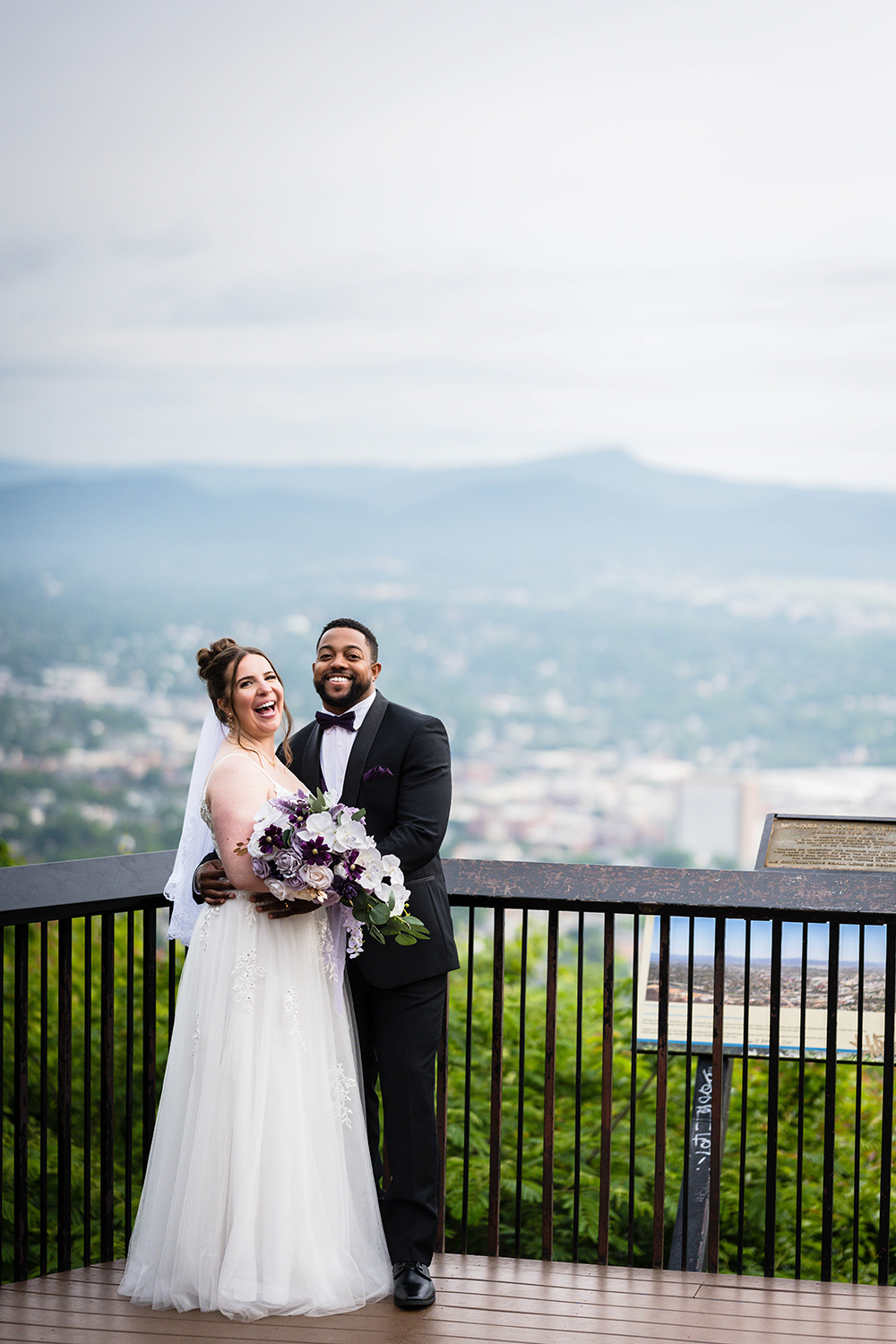 A couple wearing wedding attire stand together on their elopement day at an overlook at Mill Mountain Park. 