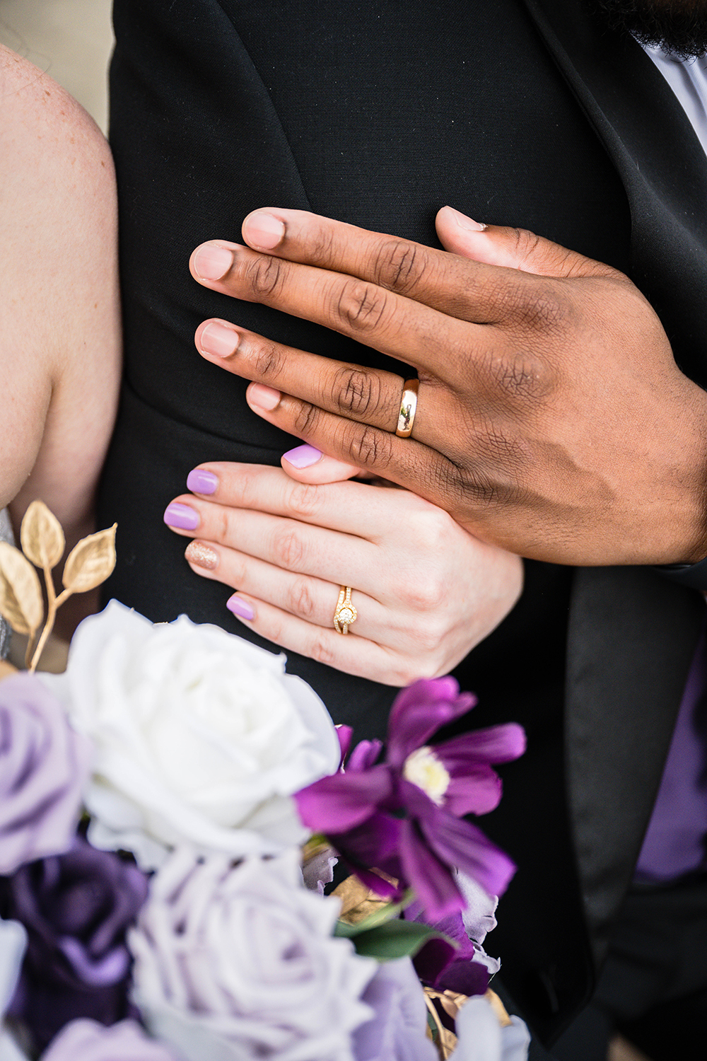 A bride wraps her arm around the groom's arm and the groom places his hand just above hers. The photo shows both of their hands featuring their wedding rings and part of the bride's bouquet.