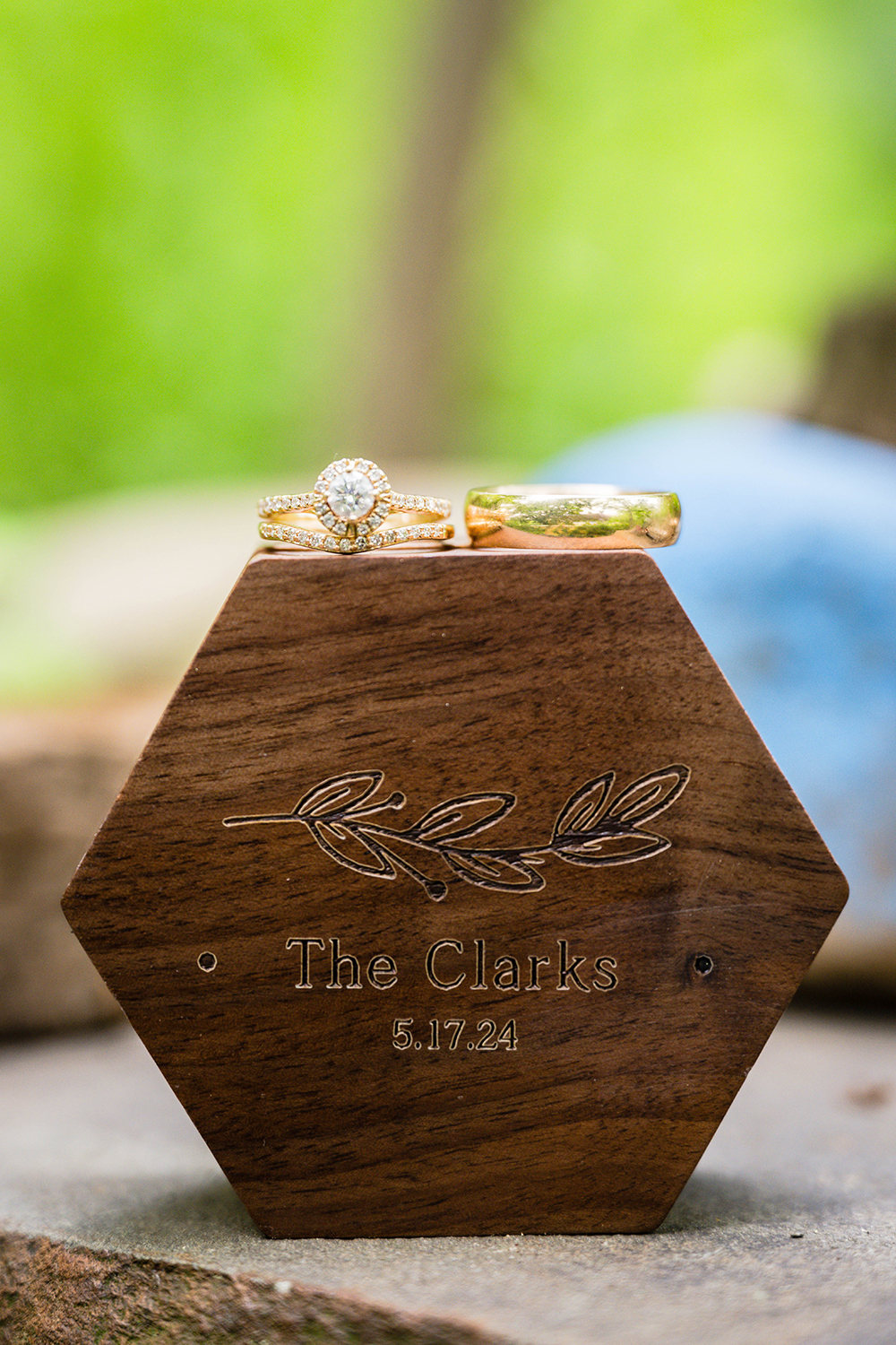 Two wedding rings sit atop of a custom ring box that has a tuft of plant and writing inscribed on the box. The writing reads "The Clarks" followed by the date of their wedding "5.17.24".