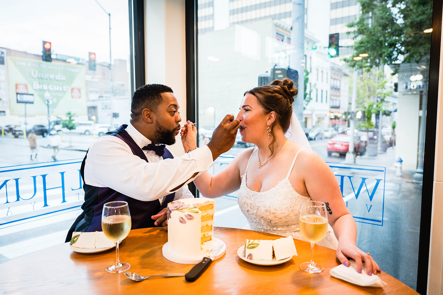 A wedding couple sits and feeds one another. They each take a spoonful of their slice of cake to feed the other. They look at one another intently.